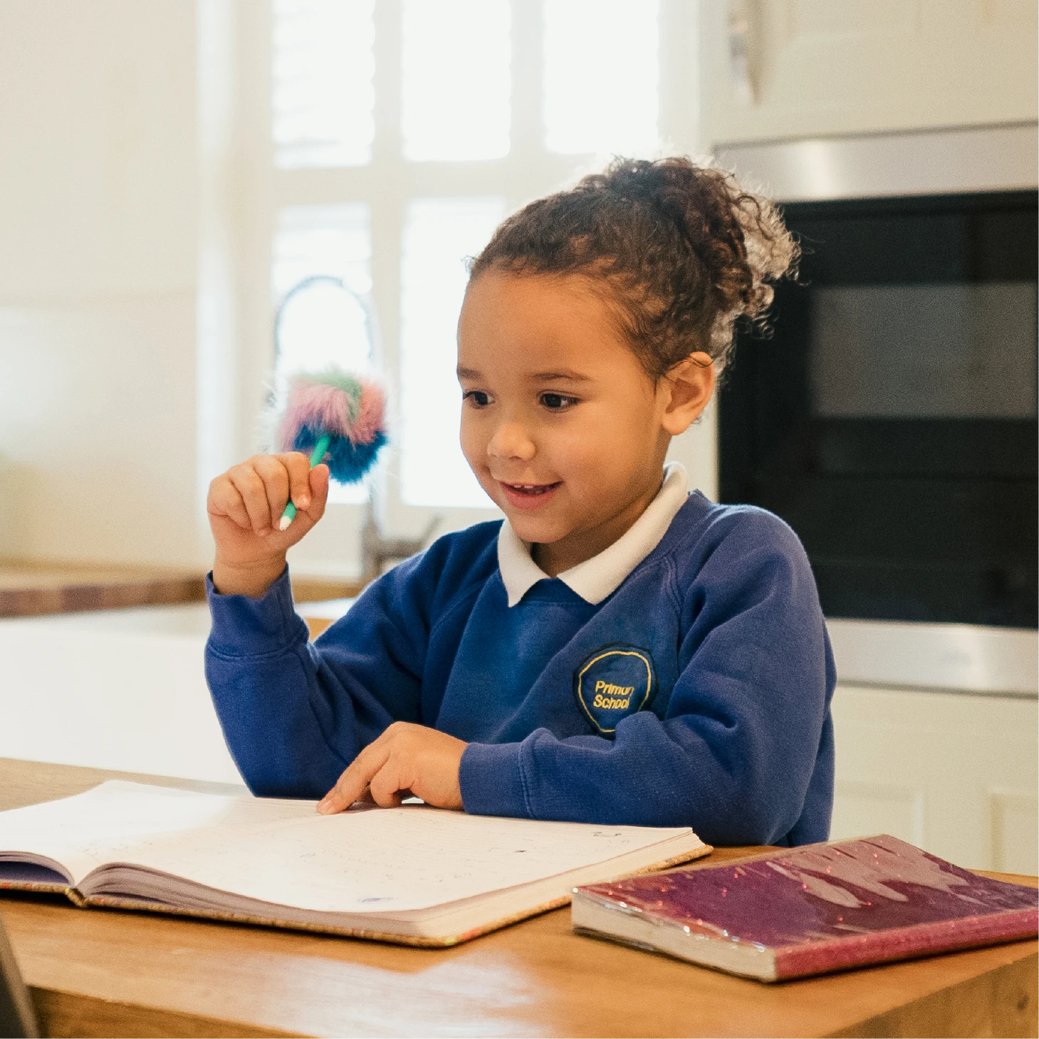 Young pupil in a blue school jumper sits at a table with an open exercise book, holding a colourful pen and smiling.