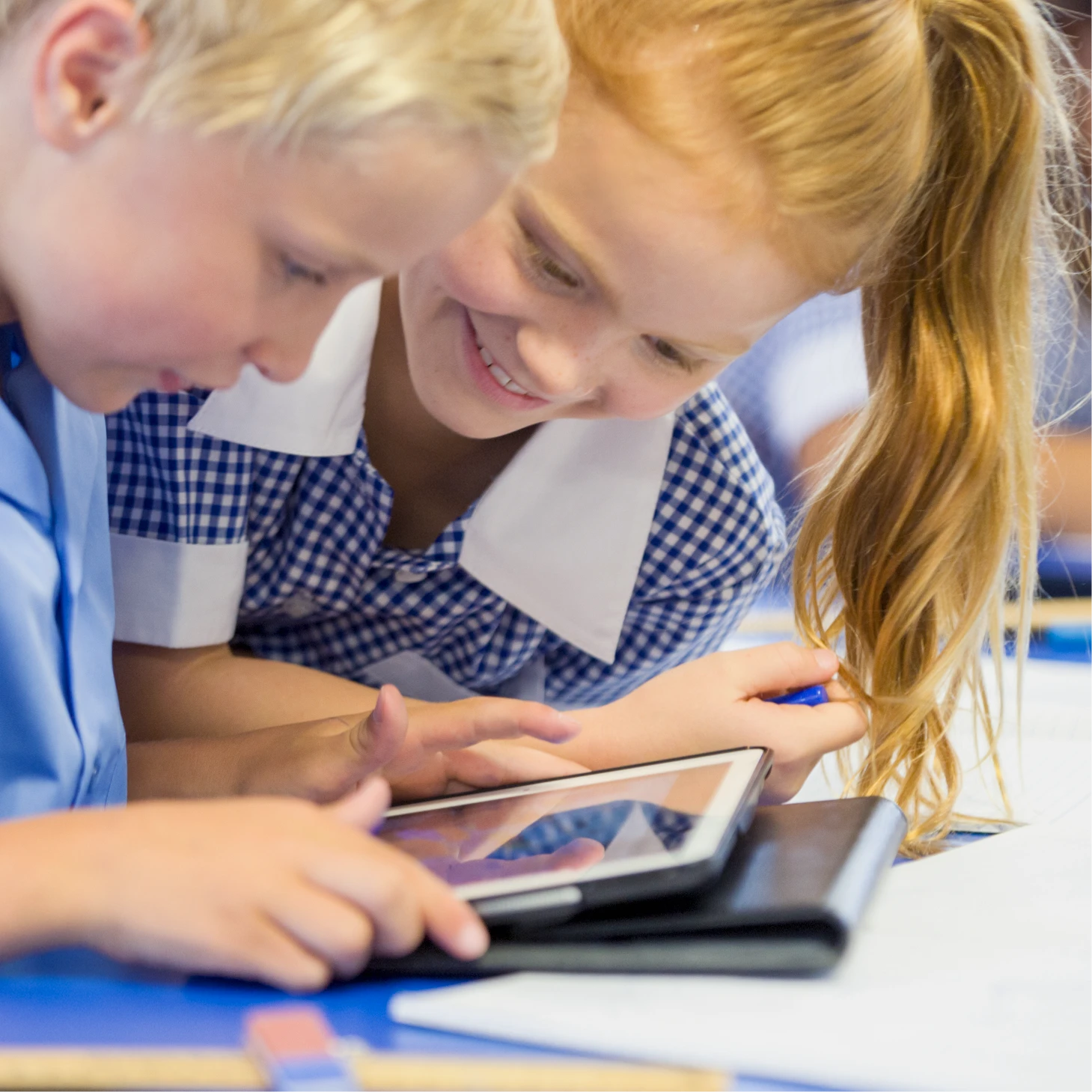 Two primary school pupils use a tablet together, looking closely at the screen during a classroom activity.