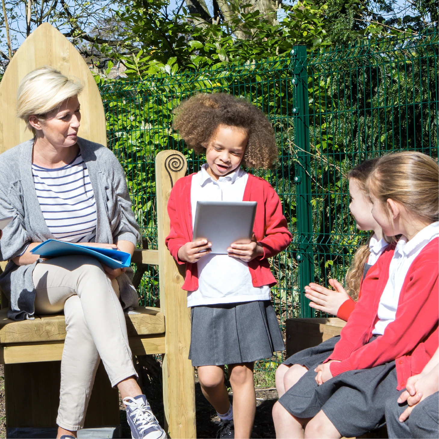 A child reading from an electronic tablet outdoors to her peers and teacher.