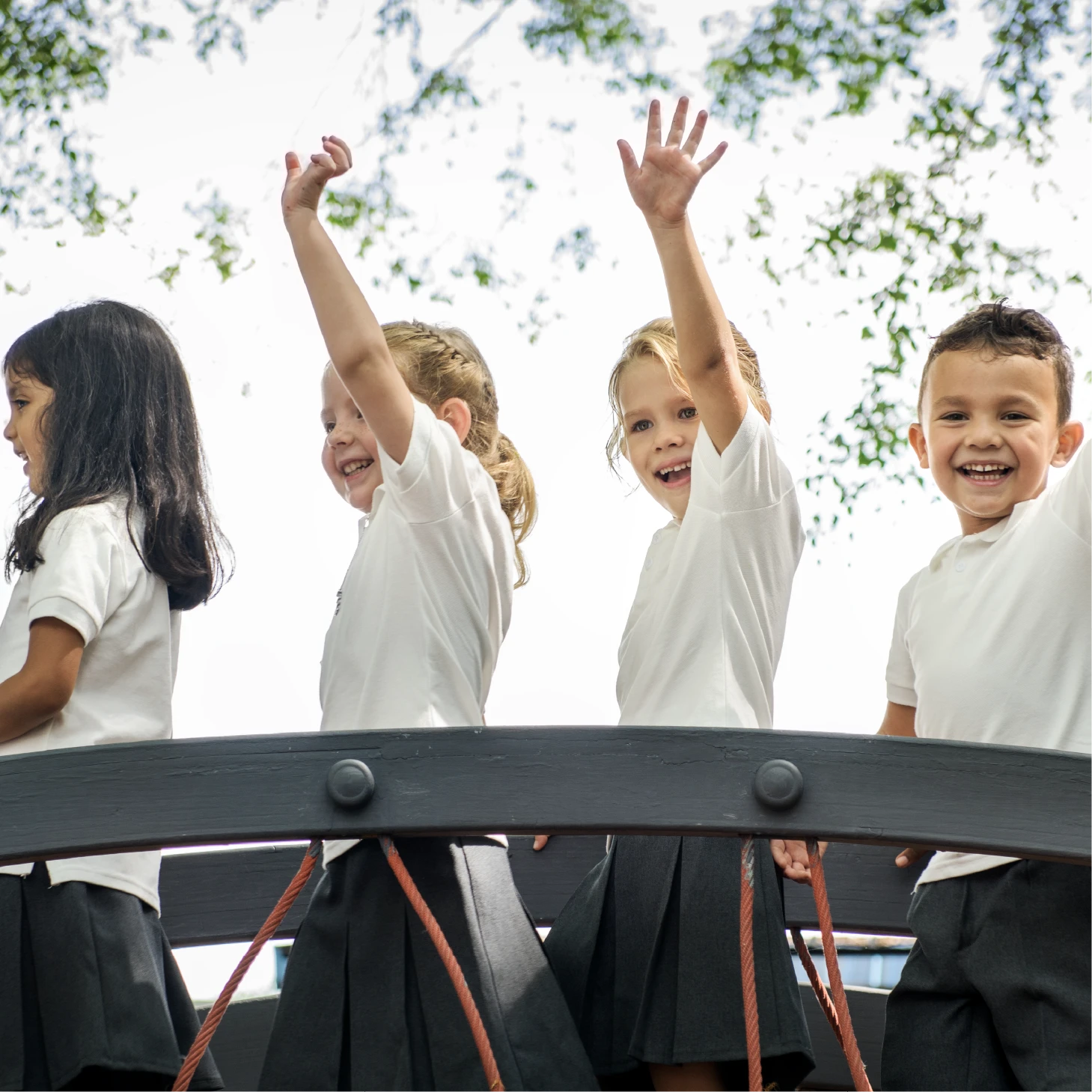Four primary school children in school uniform stand on a playground bridge, smiling and waving with raised hands.