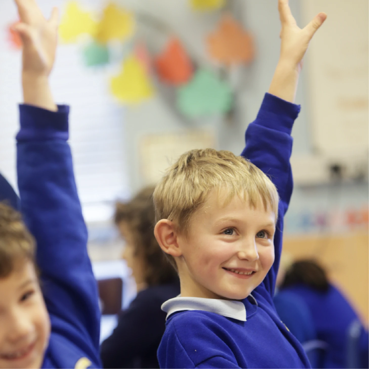 Smiling pupil in a blue school jumper raising a hand to answer a question in class.