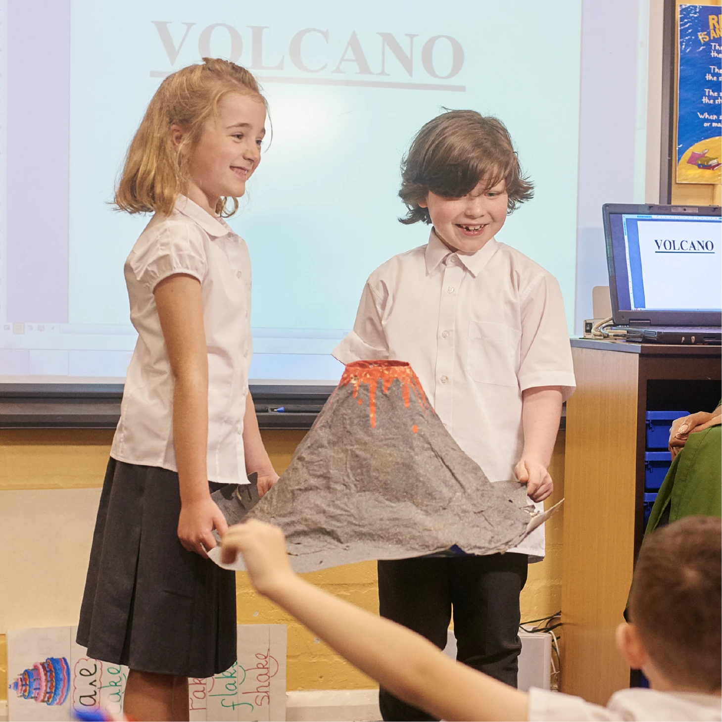 Two pupils present a model volcano to their class during a geography lesson, with a volcano presentation displayed on the interactive whiteboard behind them.