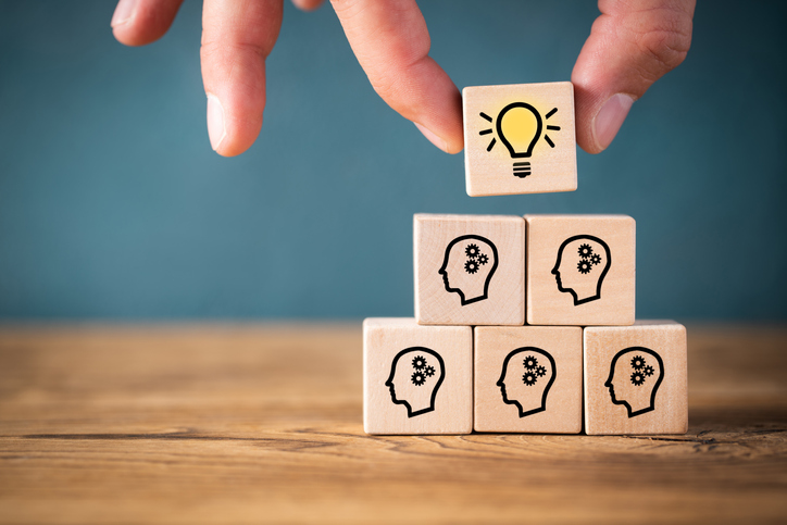 Pyramid of wooden blocks with images of heads and a lightbulb on the top one with a person placing the final cube on the pyramid.