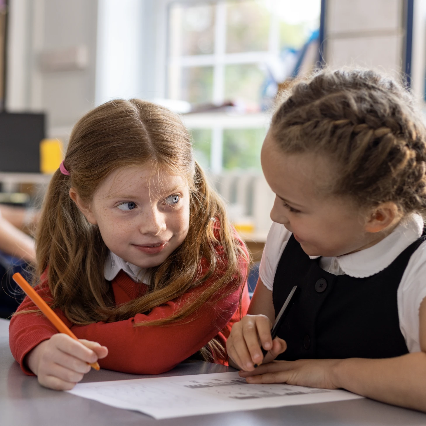 Two children sitting together at a desk, talking while working on a worksheet.
