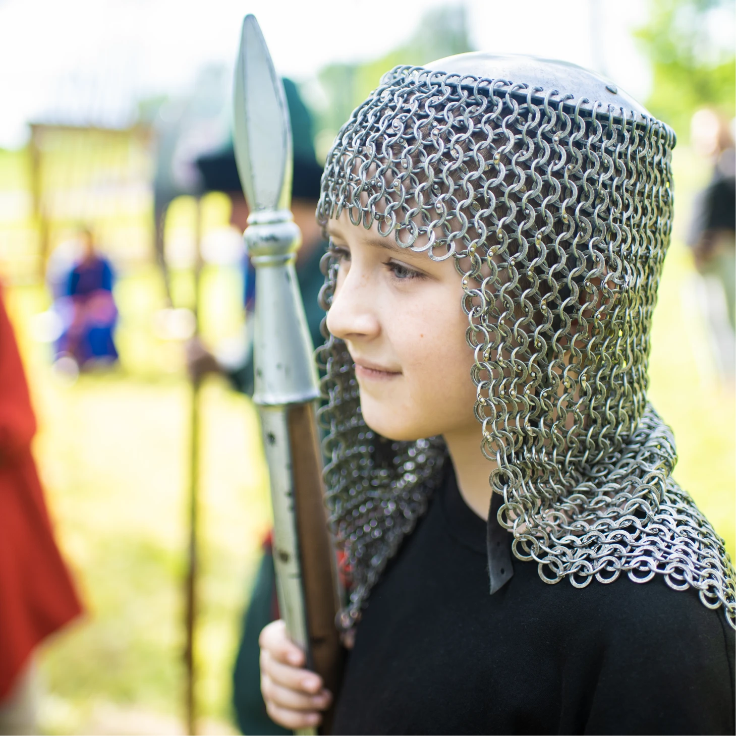 A child wearing a chainmail helmet and holding a spear at a historical reenactment.