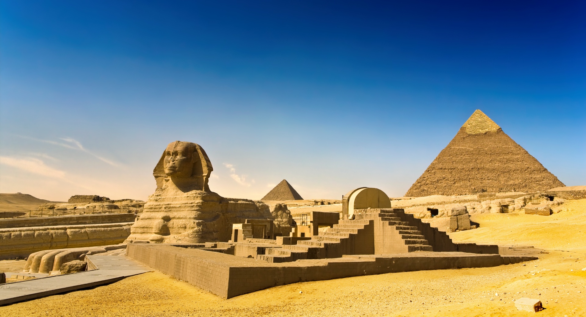 The Great Sphinx of Giza stands in the Egyptian desert with the pyramids of Giza visible in the background beneath a clear blue sky.