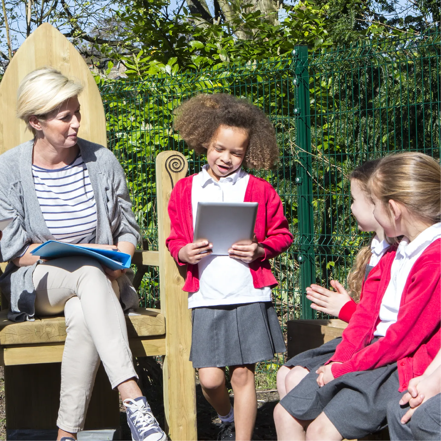 Schoolgirl reading from a tablet to classmates outdoors.