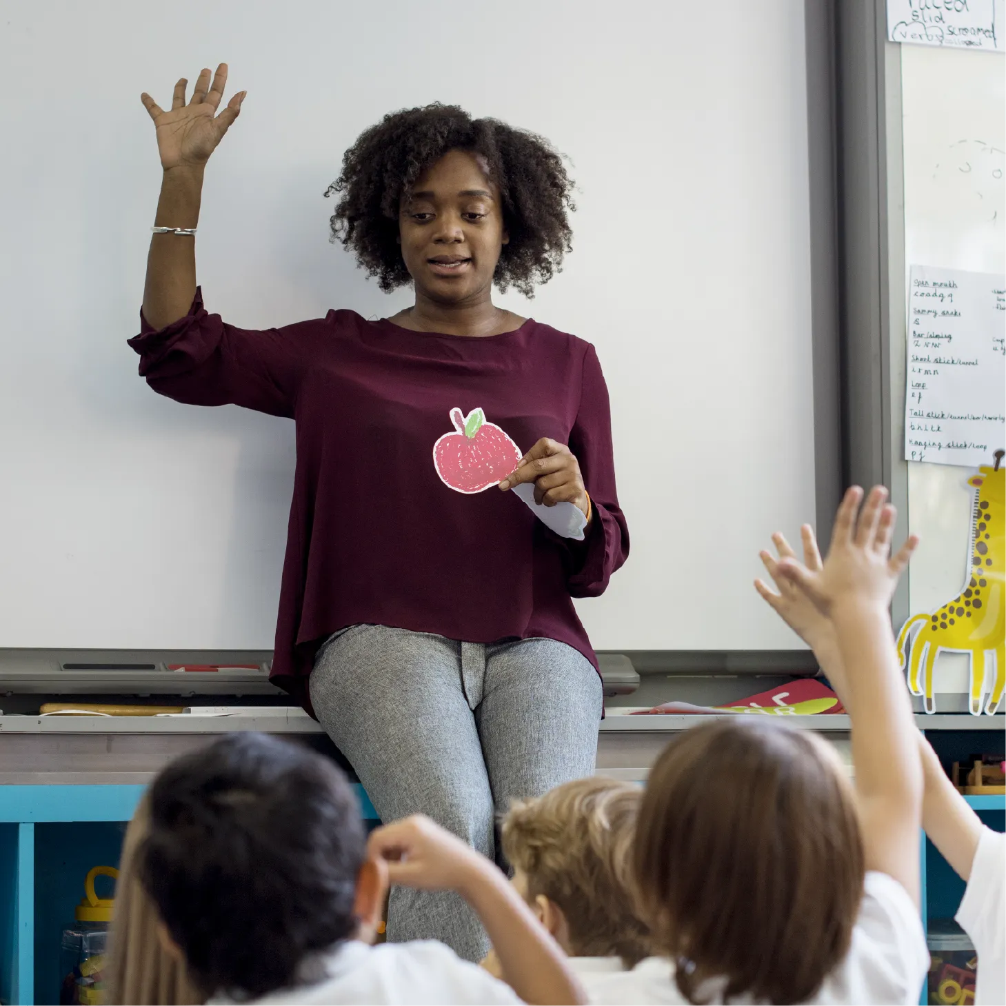 A teacher with her hand up, holding the picture of an apple, waiting for the children.