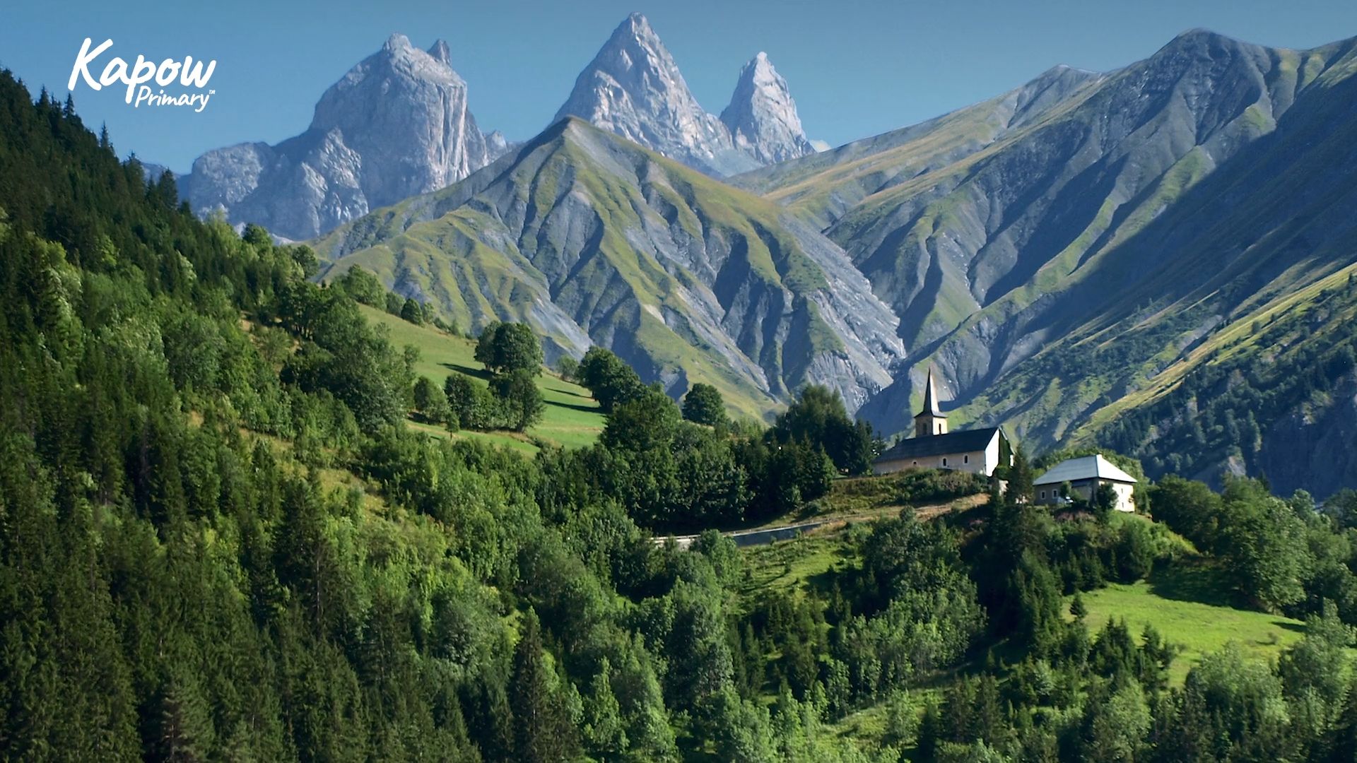 A small village sits among green hills beneath tall mountain peaks.