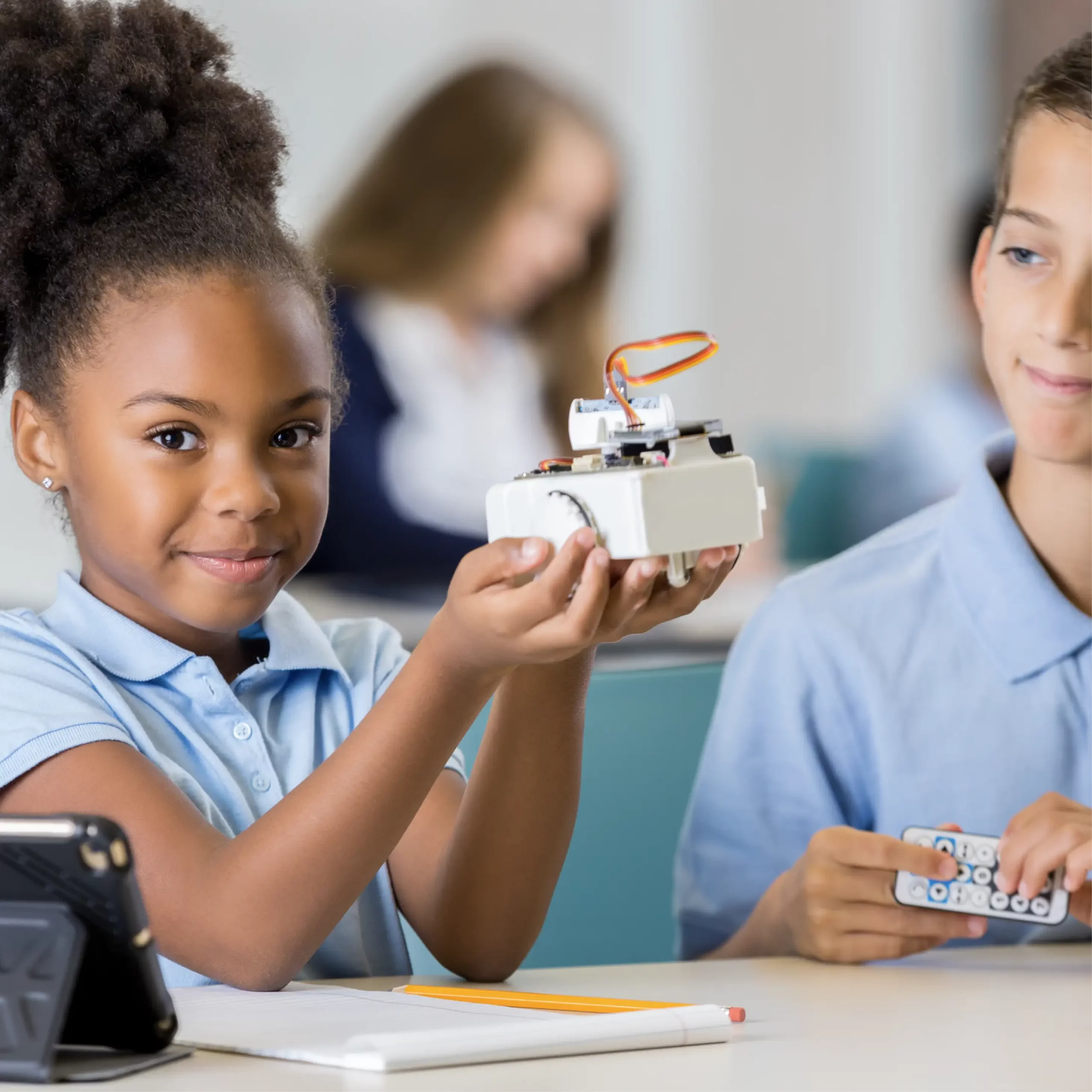 A happy girl shows an electronic item in a classroom, as a boy next to her holds the remote for it.