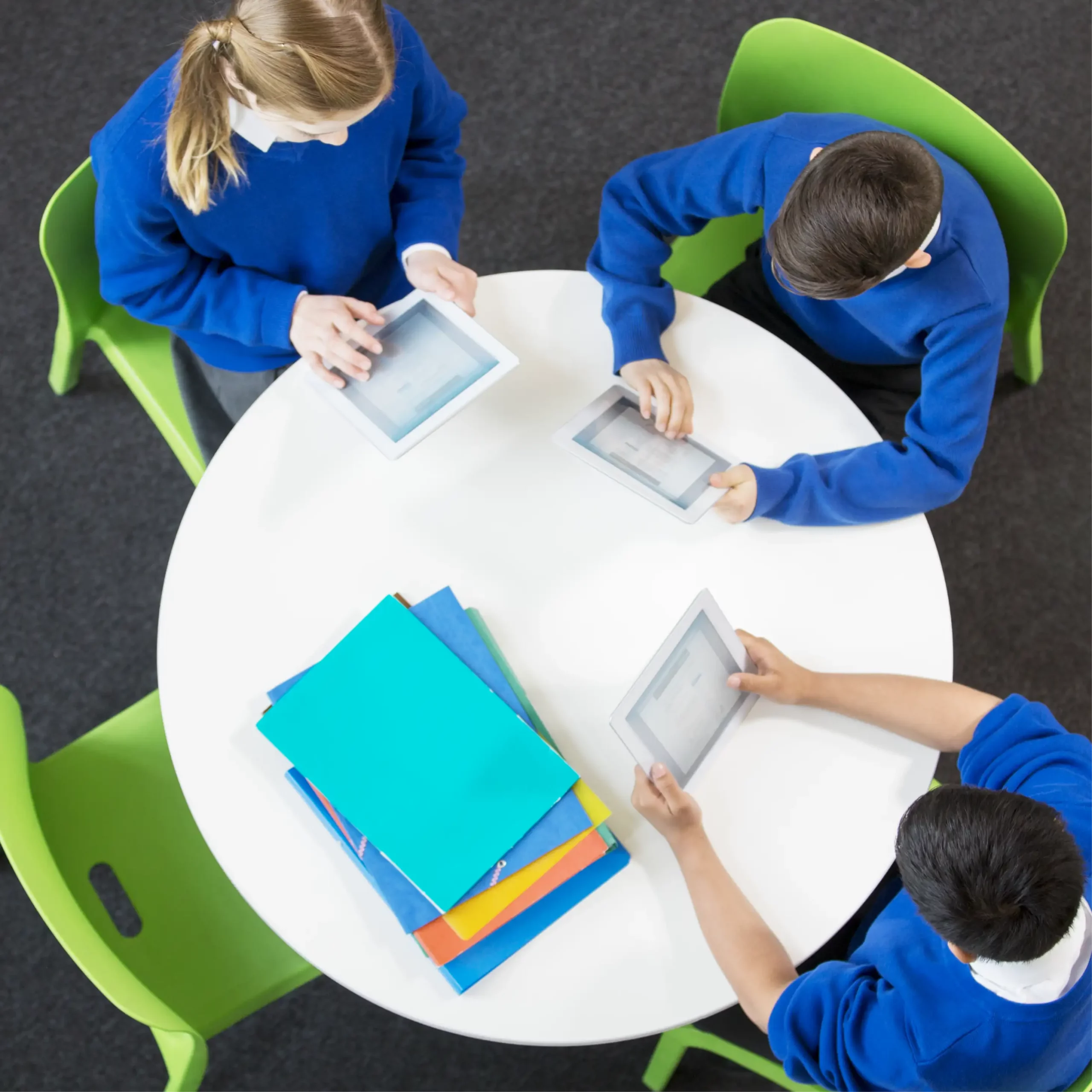 A birds eye view of three pupils in blue uniform sat around a table looking at tablets, with a pile of colourful paper on the table.