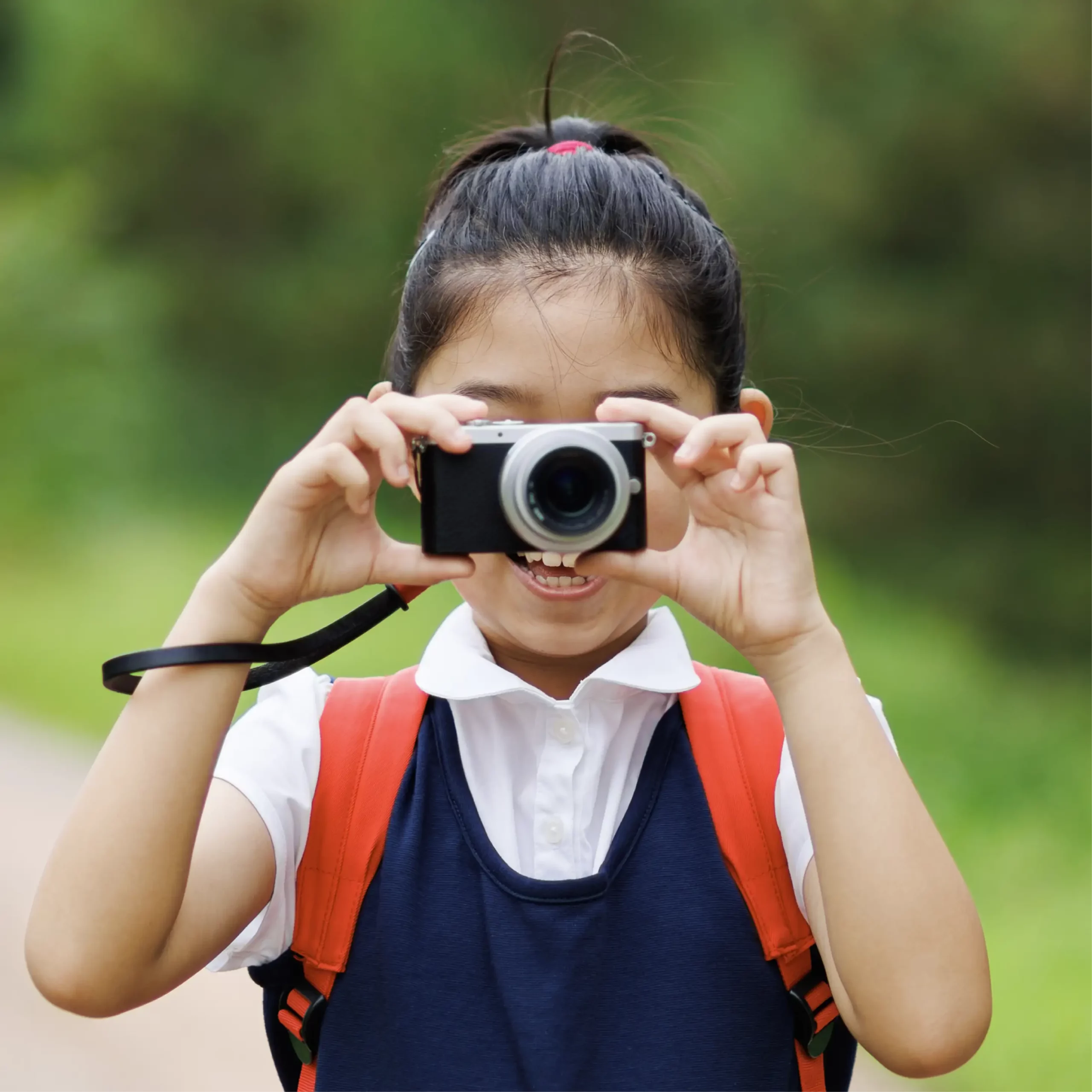 A happy girl wearing school uniform and a red backpack holds a camera in front of her face.