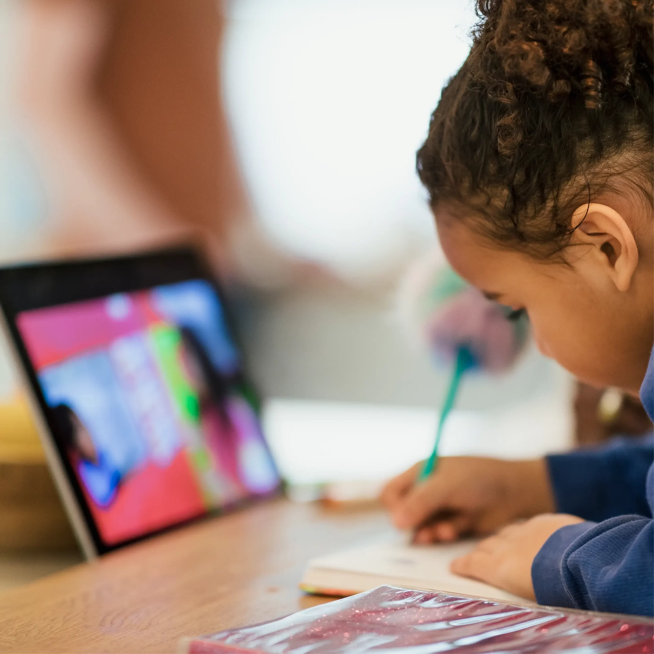 A girl writes in a notebook with a tablet in front of her.