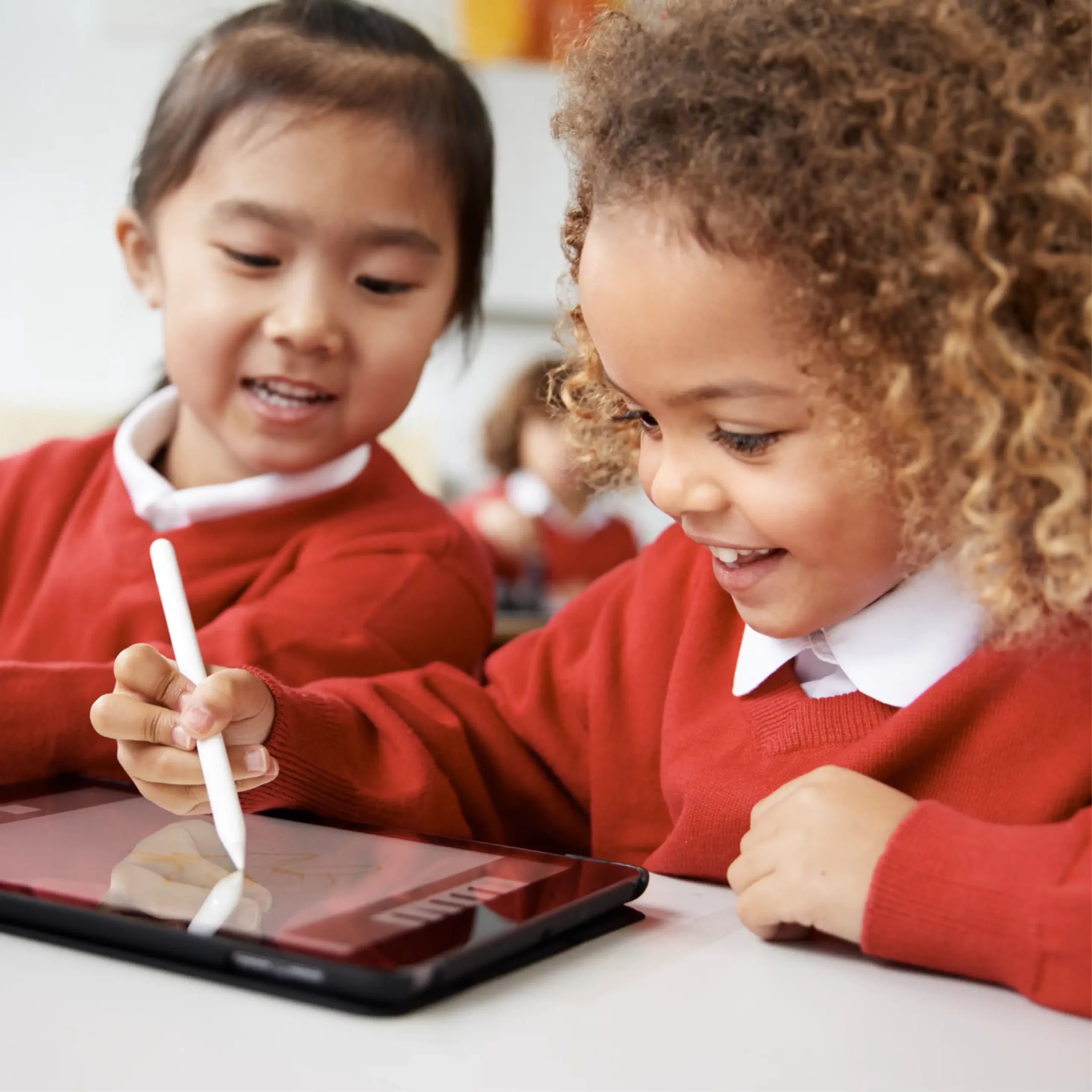 An excited girl wearing red school uniform draws on a tablet using a stylus as another girl watches her.