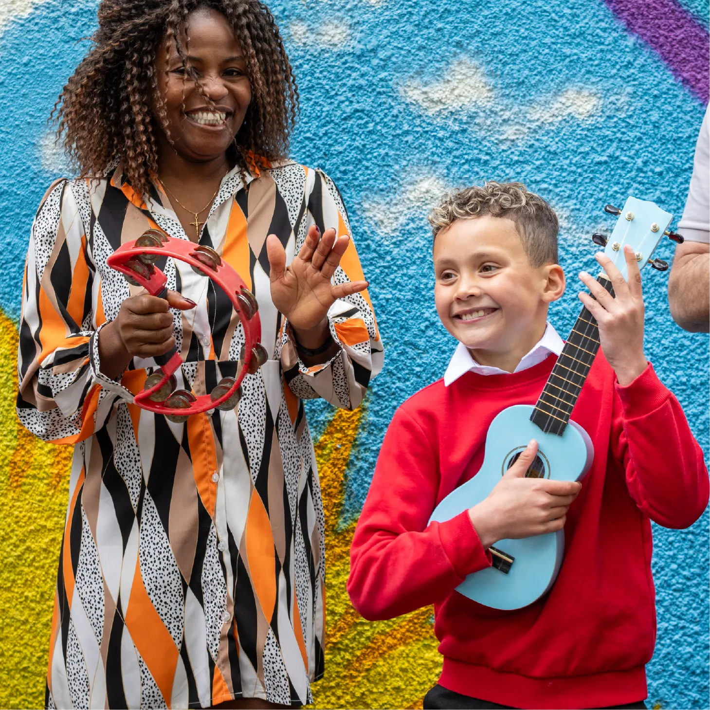 Smiling teacher and primary school pupil making music outdoors with a tambourine and a blue ukulele.