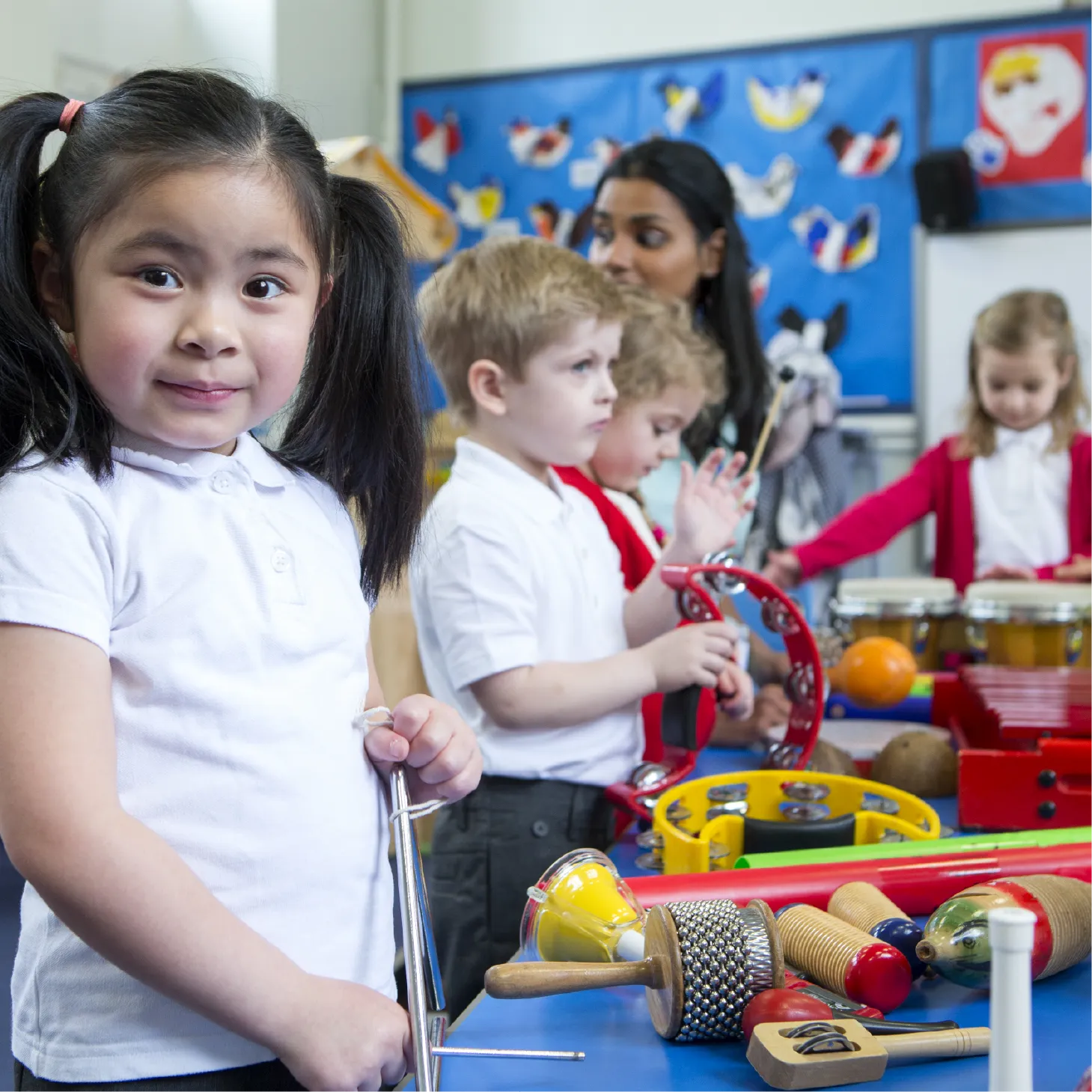 Young children in a classroom play a variety of percussion instruments, including a triangle, tambourine, and bongos, supported by a teacher in the background.