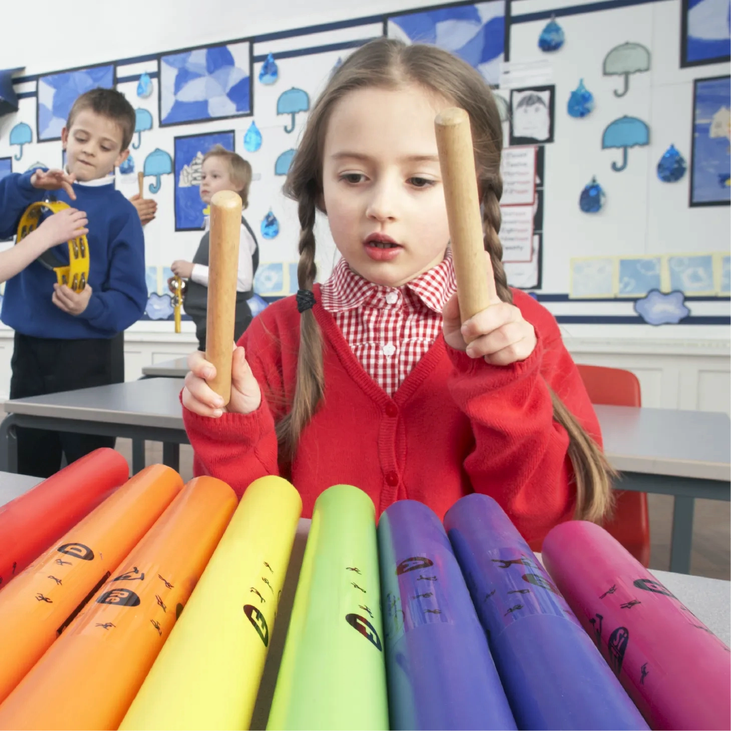 A young girl in a red school uniform plays brightly coloured Boomwhackers with wooden beaters in a classroom, while other children play tambourines in the background.