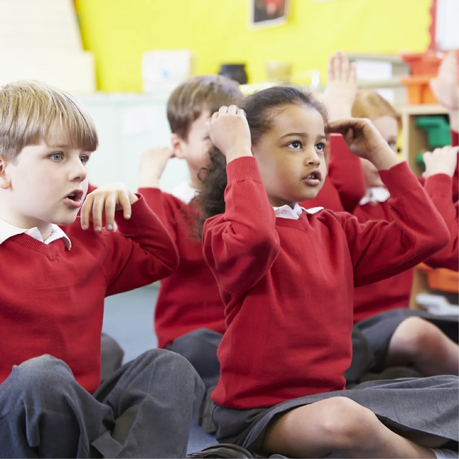 Primary school children in red uniforms sit on the floor singing and using hand actions during a classroom activity.