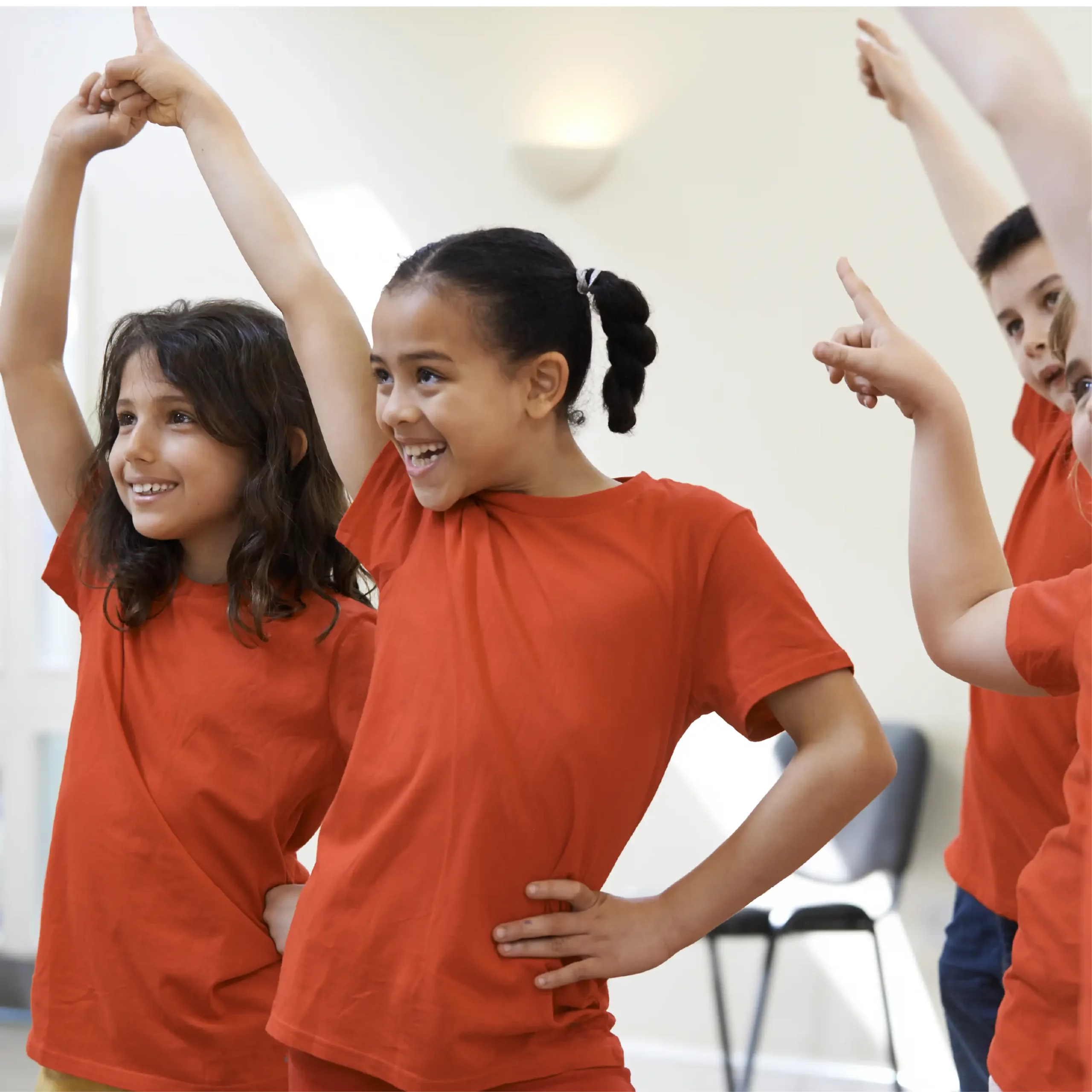 Children in red T-shirts smiling and raising one arm in the air while taking part in a group dance activity.