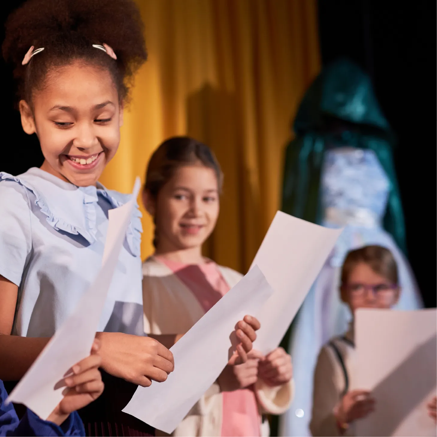 Group of children on stage holding scripts, smiling as they rehearse lines together.