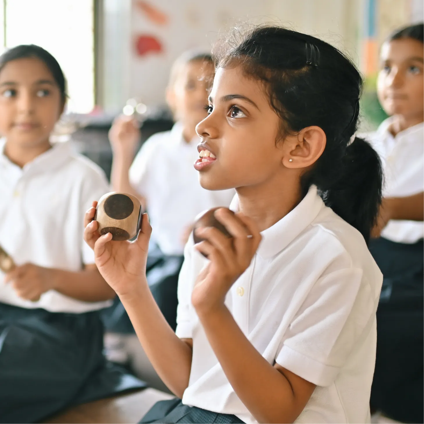 Primary school children in white uniforms playing handheld percussion instruments in a classroom.