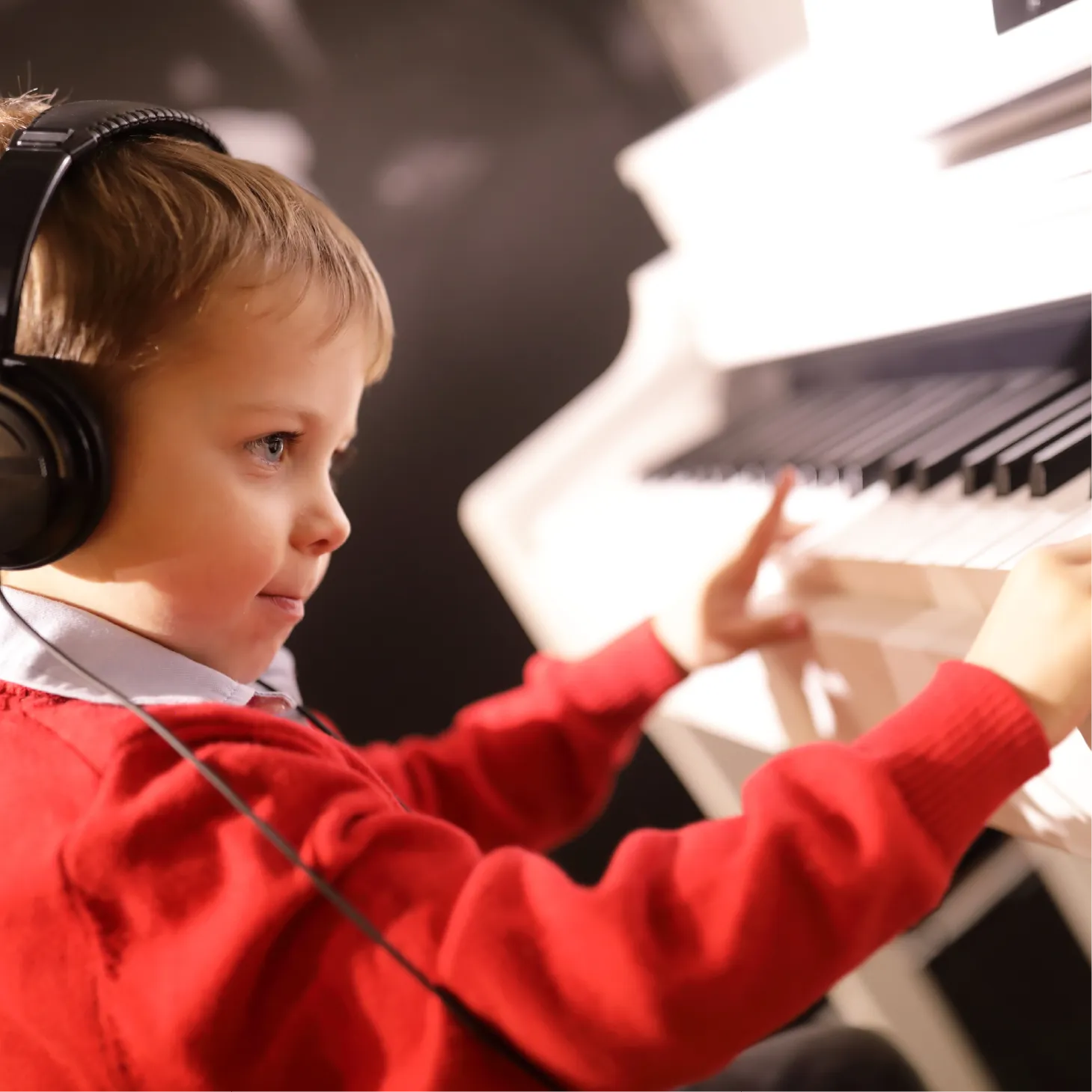 A young boy in a red jumper wearing headphones plays a white keyboard piano with concentration.
