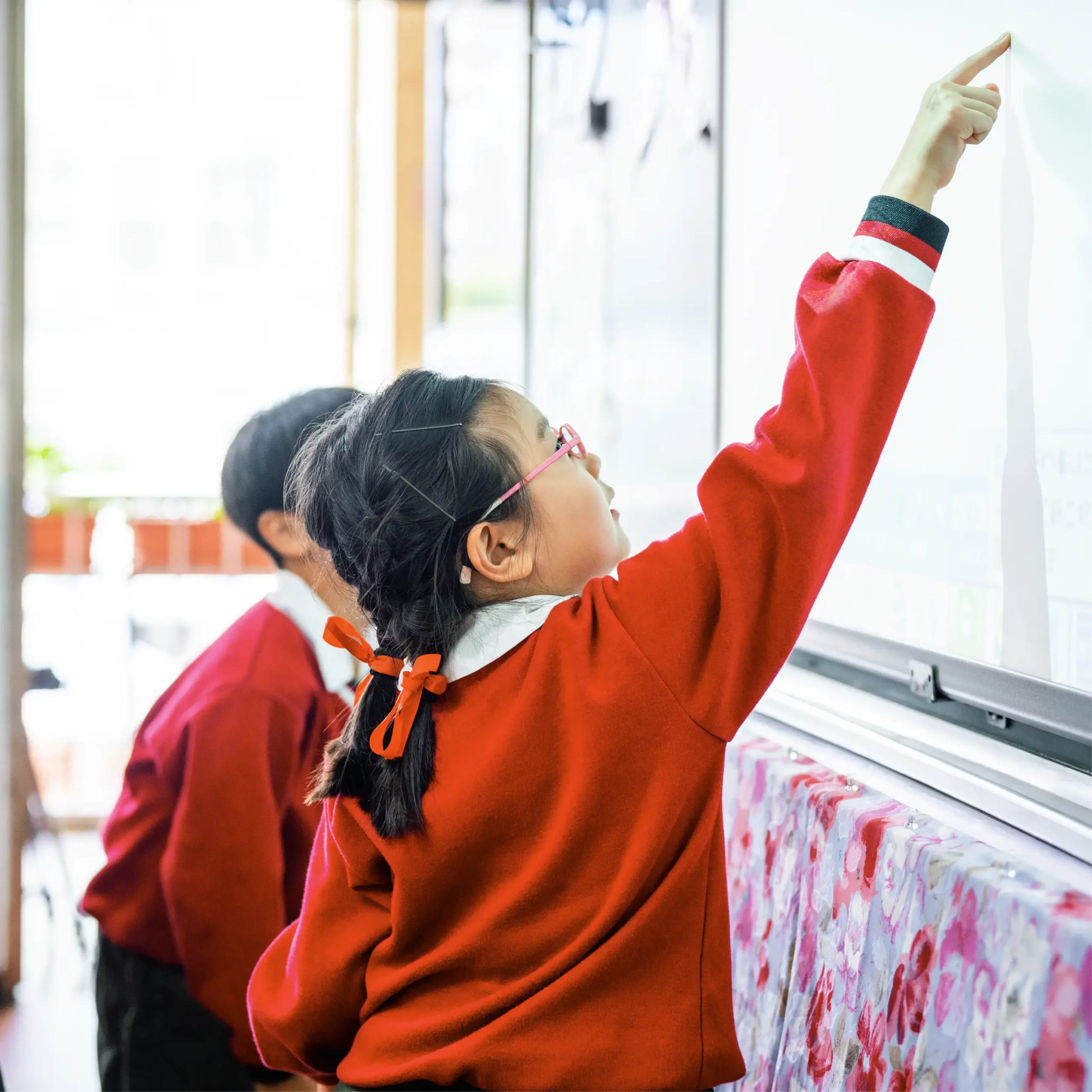 Child in a red jumper with glasses and braided hair pointing up at a classroom whiteboard while another student looks on.