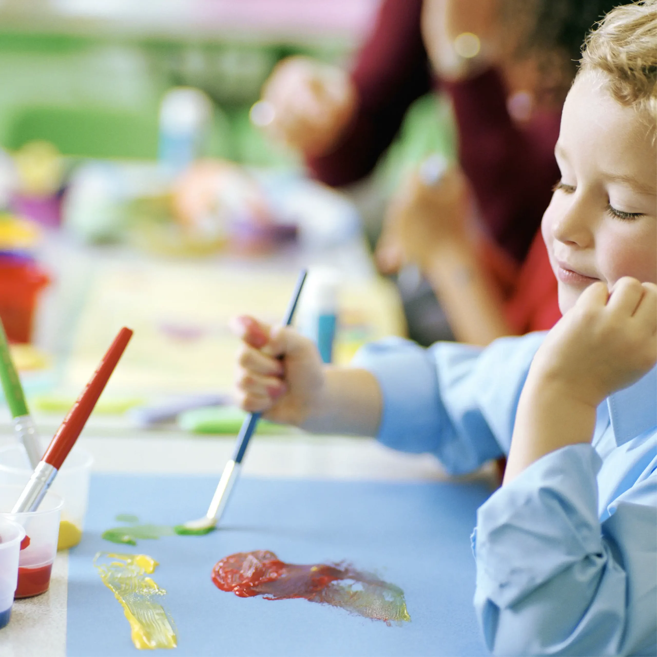 Child in blue shirt paints with red, yellow and green colours on paper during a classroom art lesson.