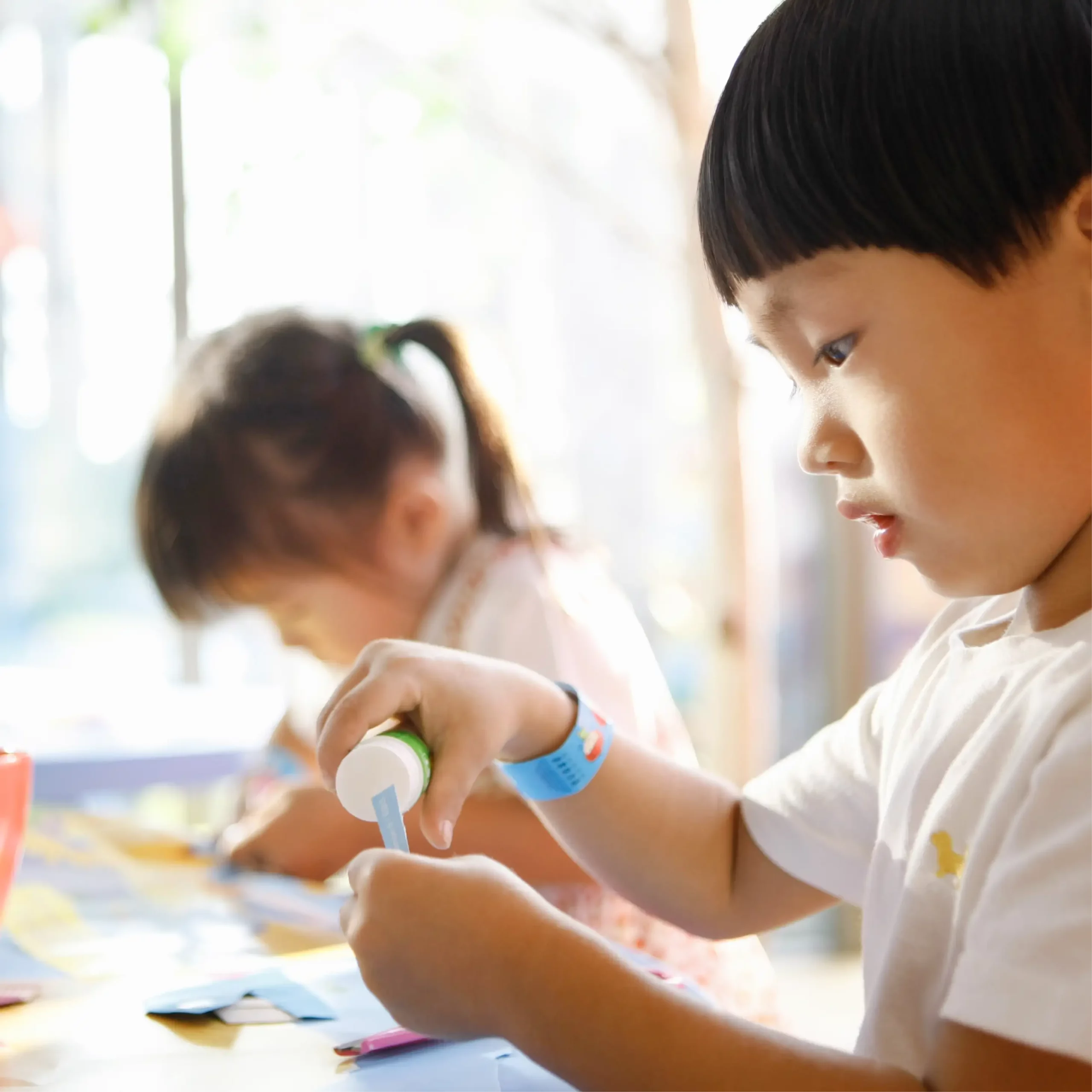 A child carefully applies glue to a piece of paper while another child in the background also works on a craft project at a table.