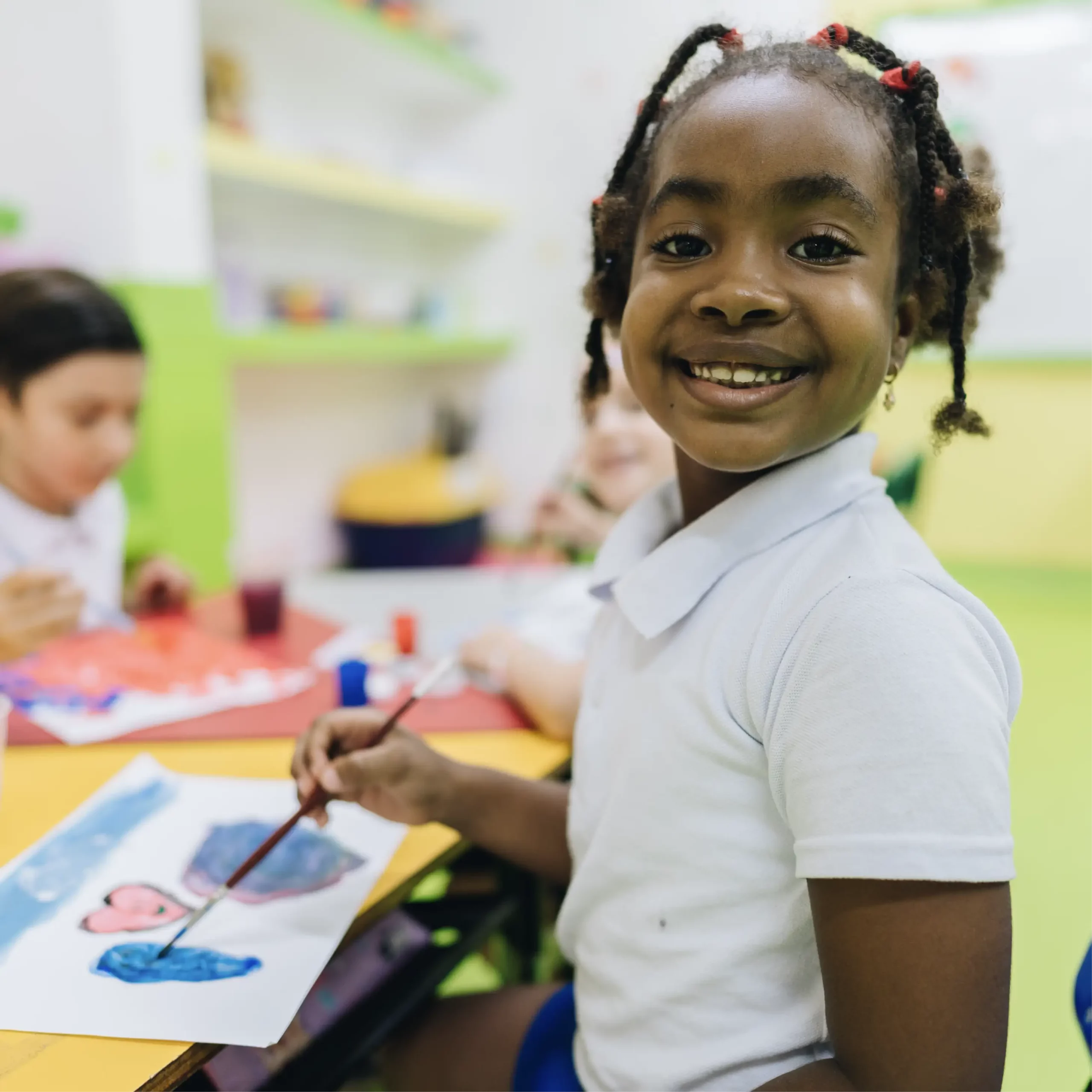 Smiling child in a white shirt paints with blue on paper at a classroom table, with other children painting in the background.