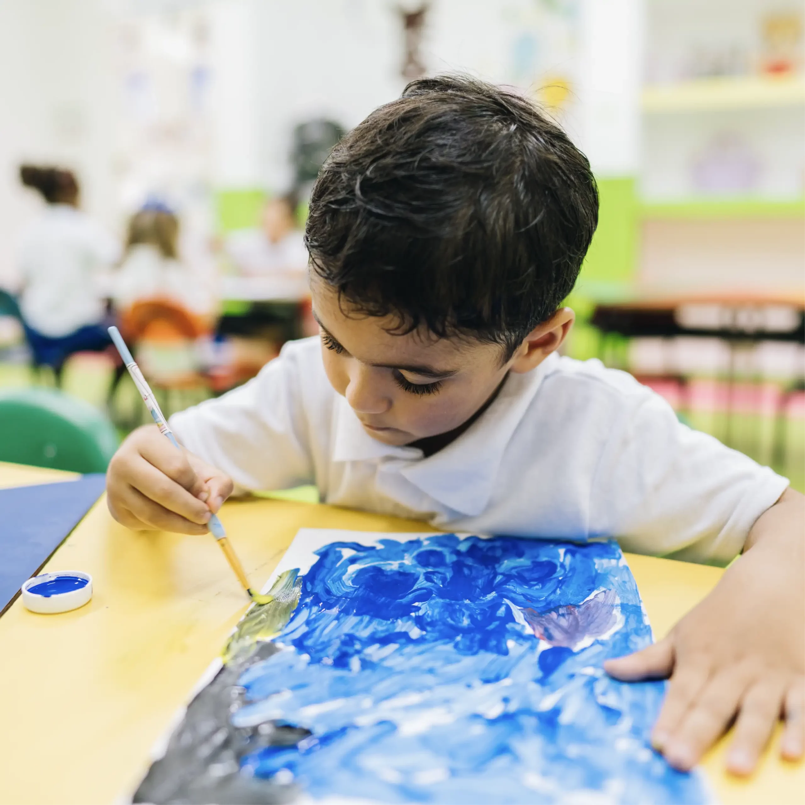 Child concentrating while painting with blue and green colours on paper during an art activity.