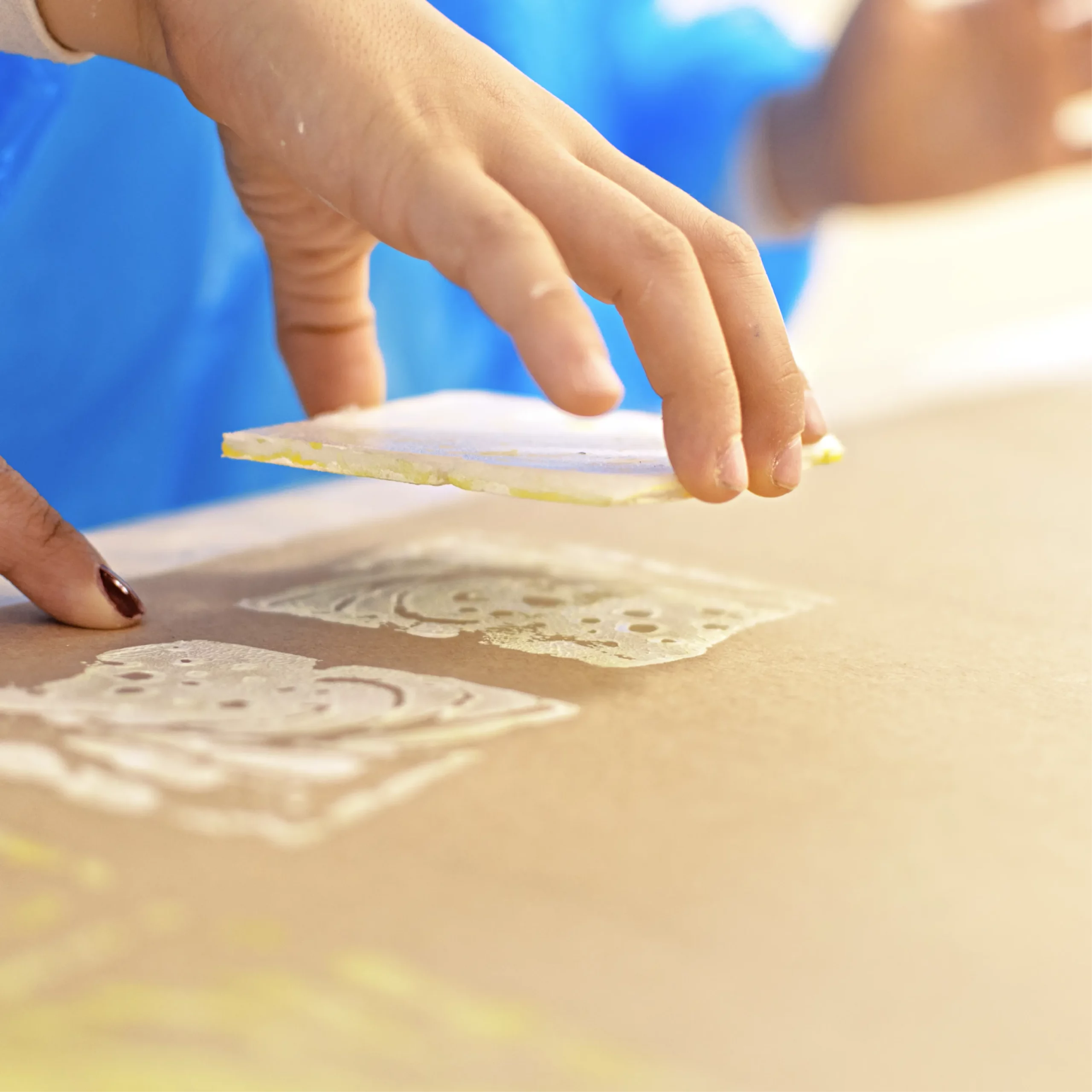 Child’s hand creating block prints on paper, lifting a square stamp to reveal painted decorative patterns in white and yellow.