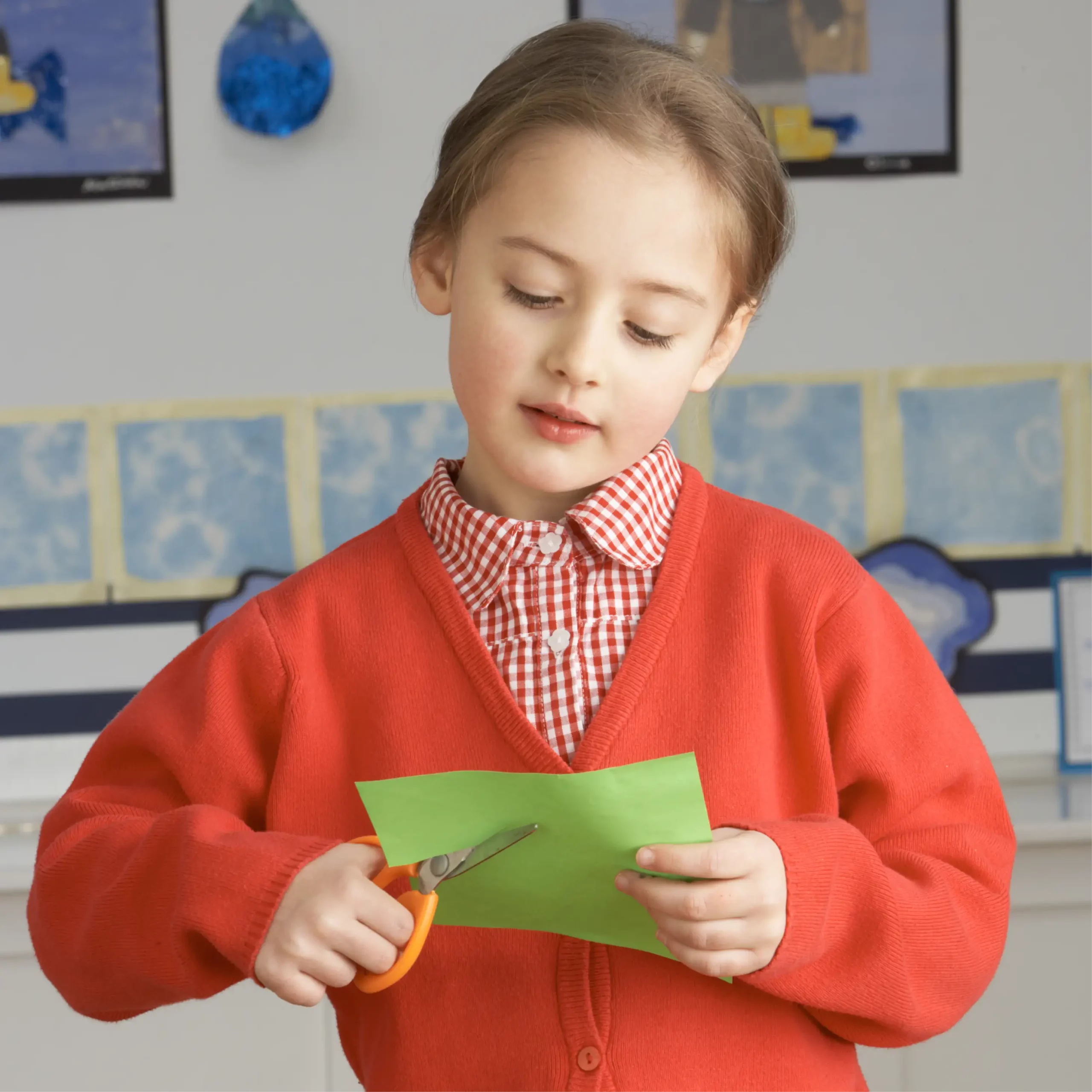 Child in red school uniform cuts green paper with scissors during a classroom craft activity.