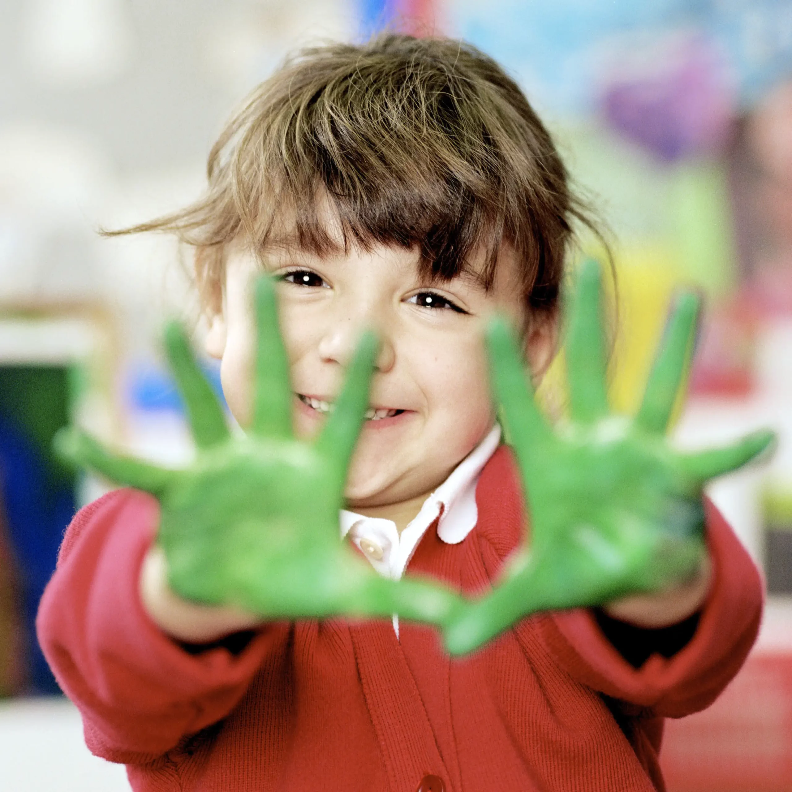 Close-up of a child in a red school jumper smiling and holding out both hands covered in bright green paint.