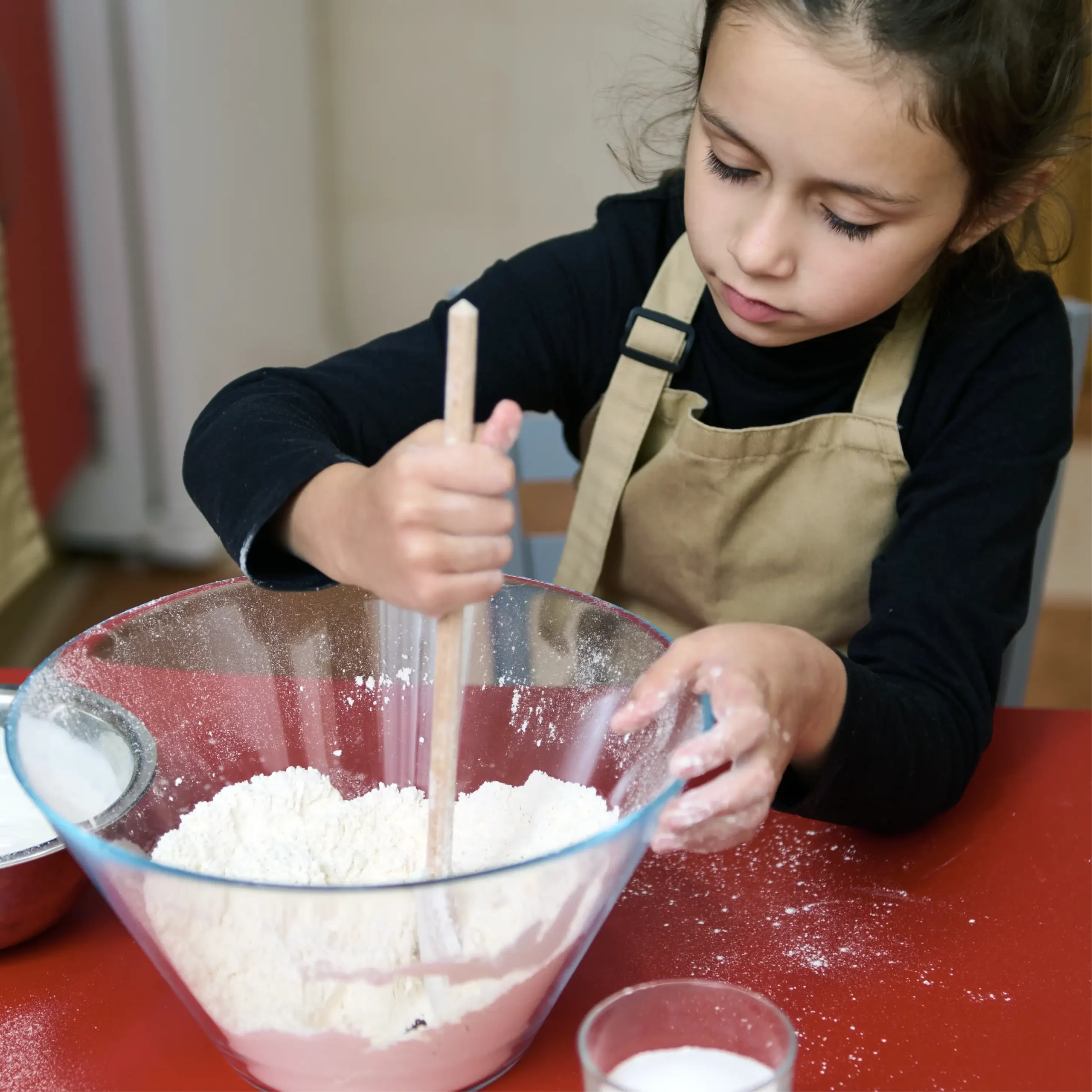 Child wearing an apron mixing flour in a large glass bowl with a wooden spoon.