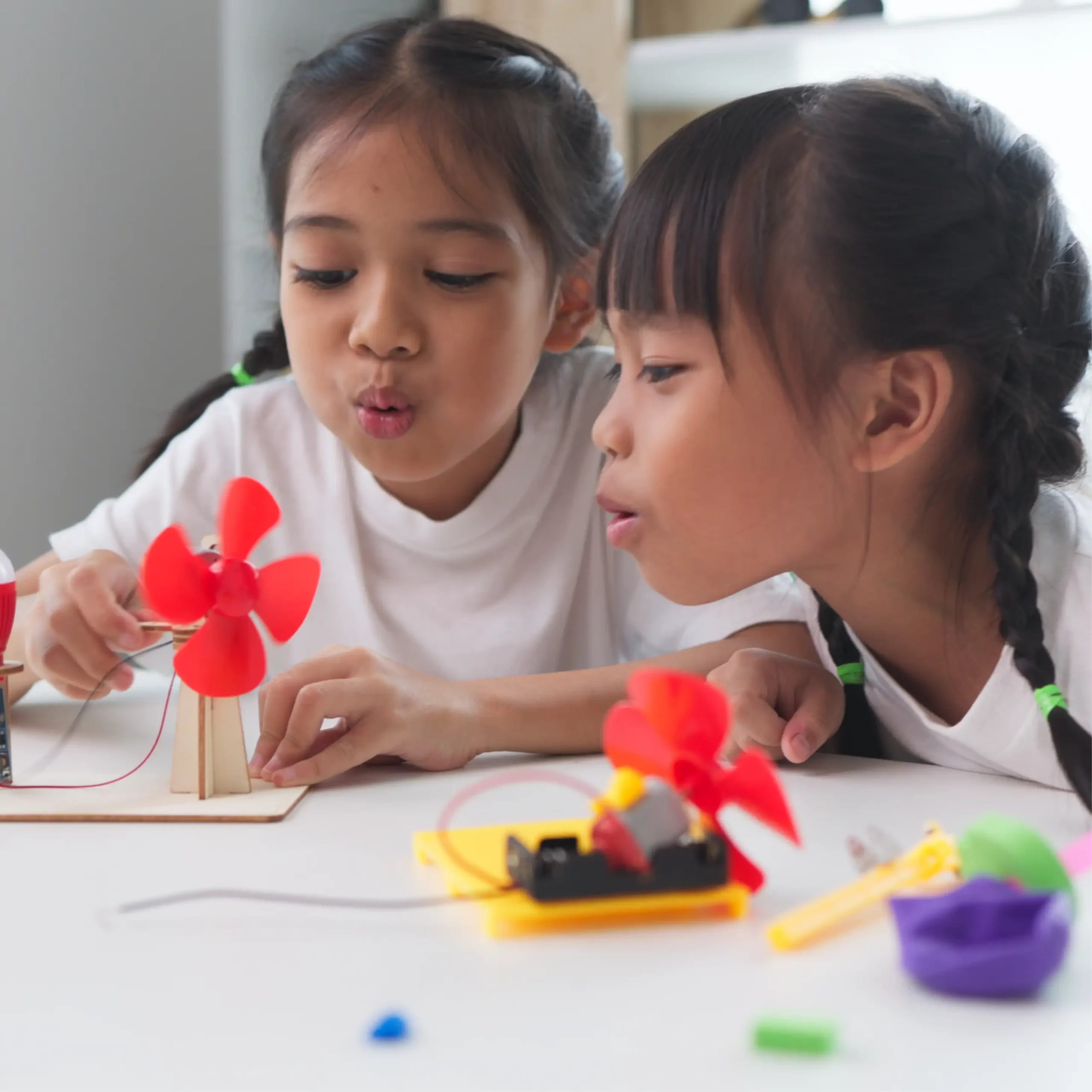 Two children test small red-bladed fan projects on a table, one powered by wires and a motor.