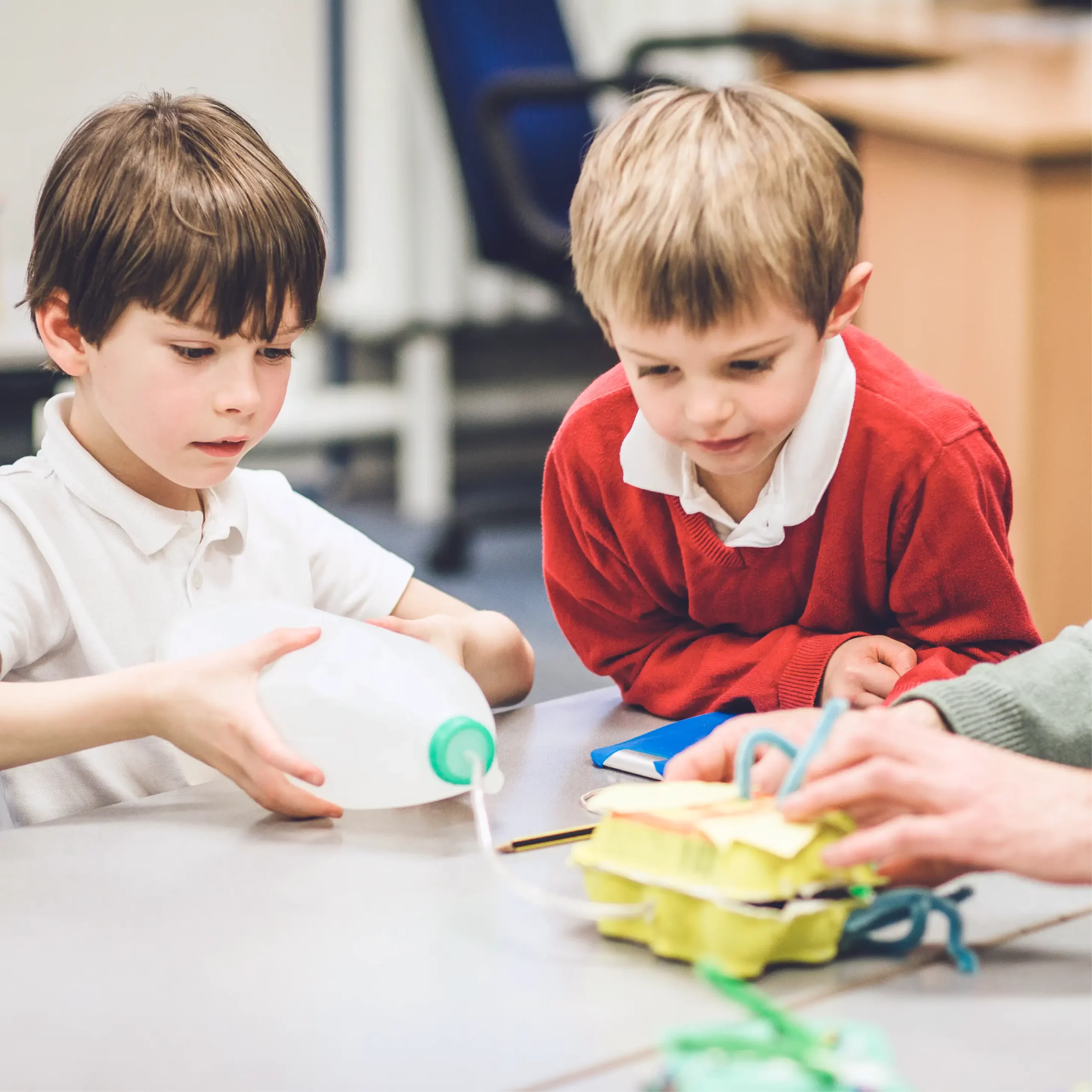 Two young boys working on a science project with recycled materials, including a plastic milk jug and egg cartons.
