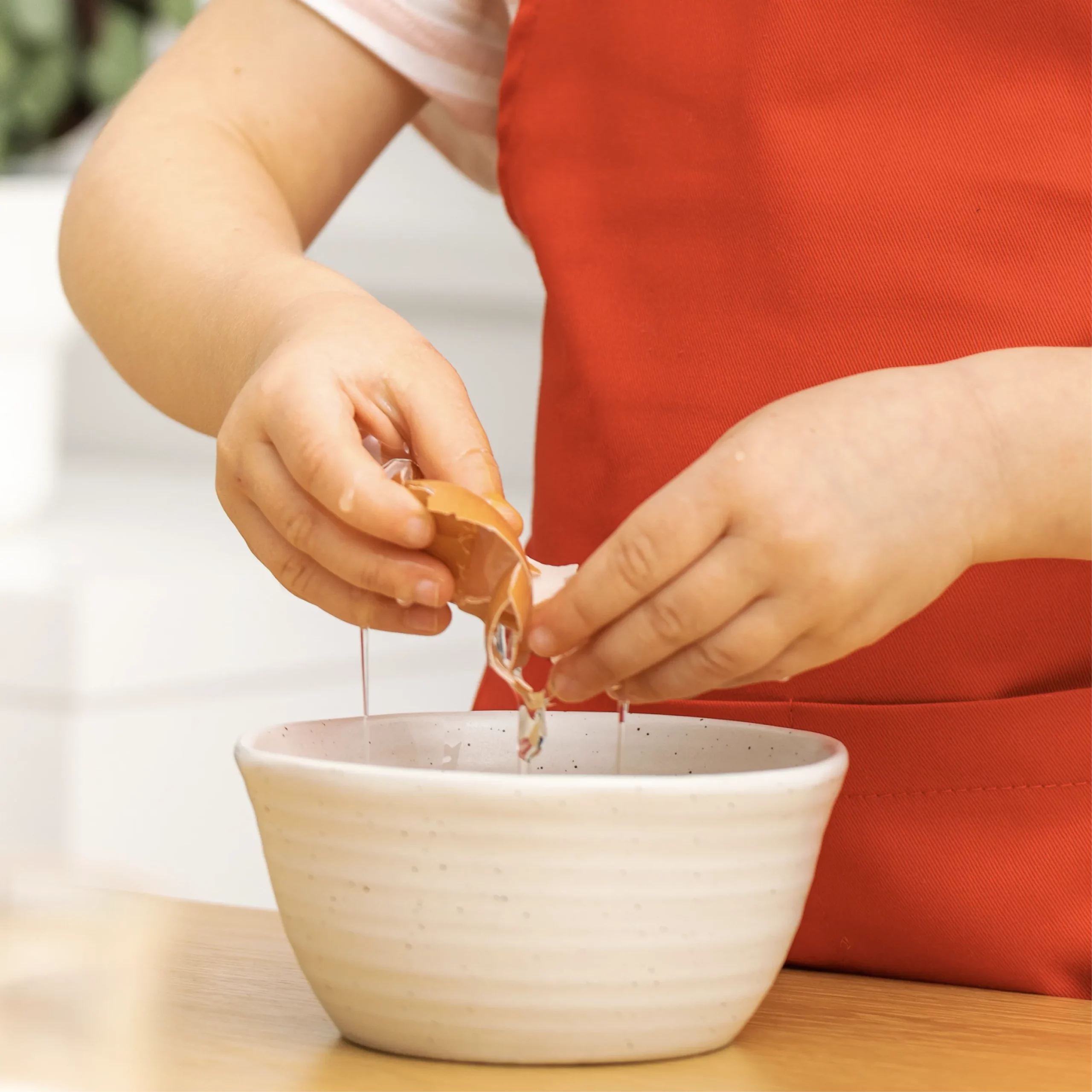 Child in a red apron cracking an egg into a white mixing bowl.