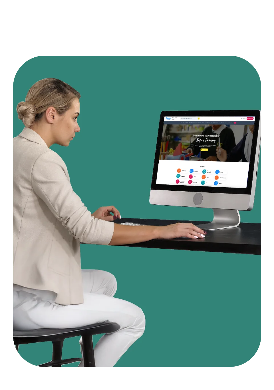 A woman sits at a desk using a desktop computer to access the Kapow Primary website for teaching resources.