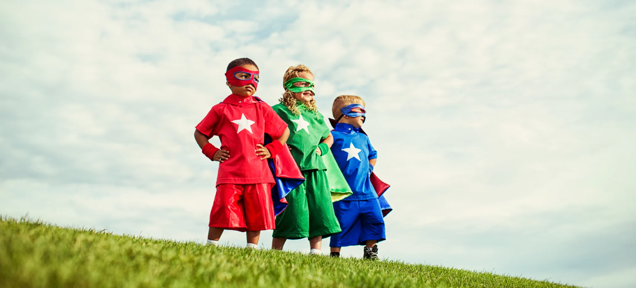 Three children wearing superhero costumes stand on a grassy hill with hands on their hips.