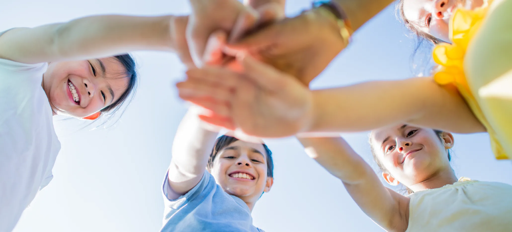 Children smiling and stacking their hands together while standing in a circle outdoors.