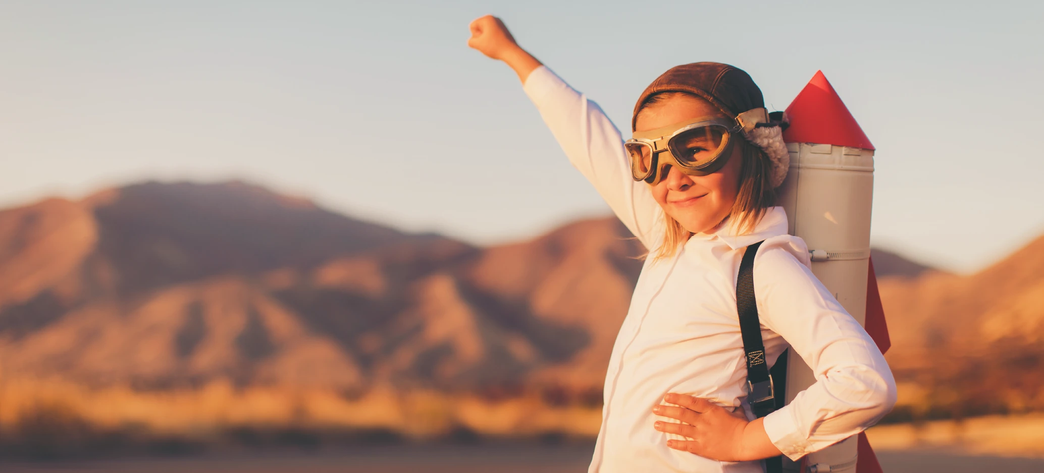 A child wearing goggles and a rocket backpack raises a fist in a superhero pose outdoors.