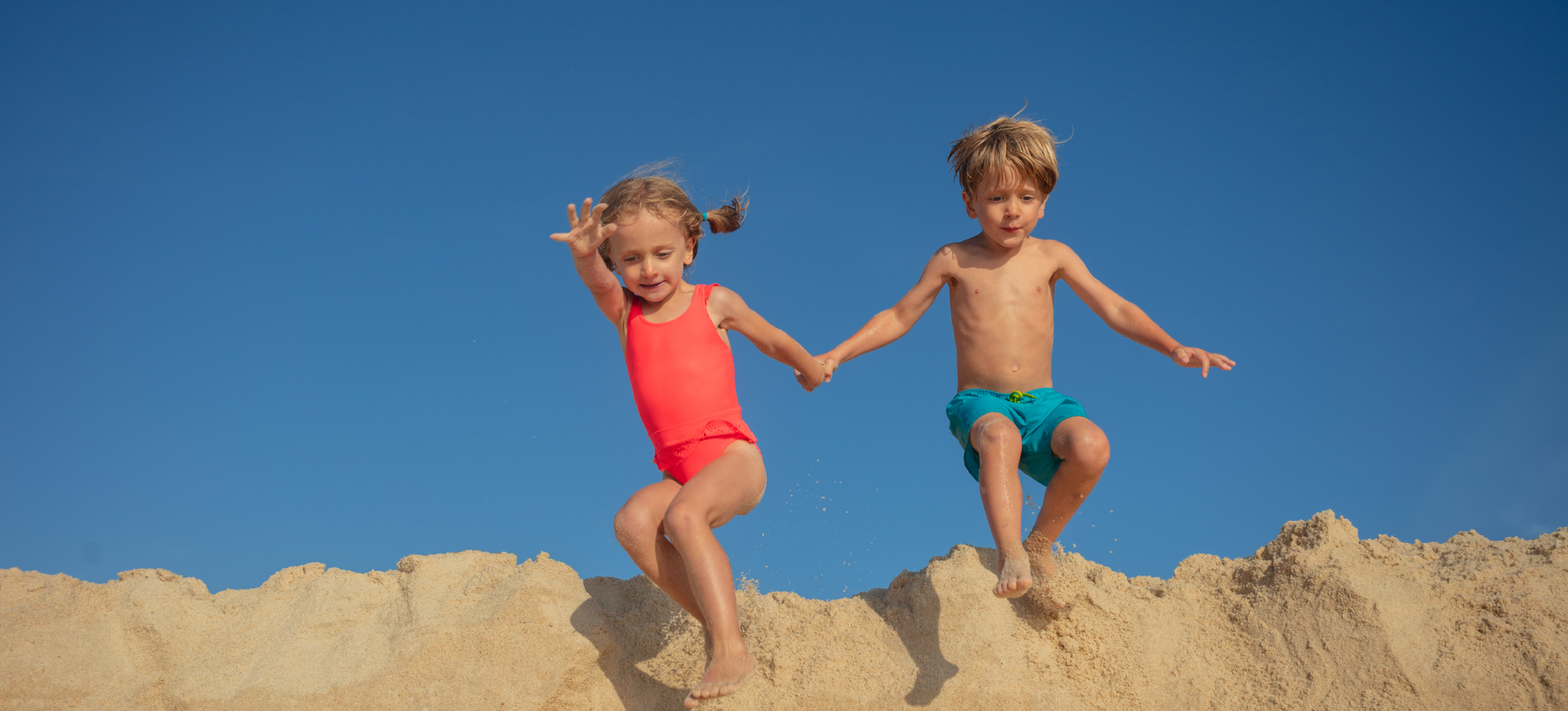 Two children holding hands as they jump down from a sandy hill under a clear blue sky.
