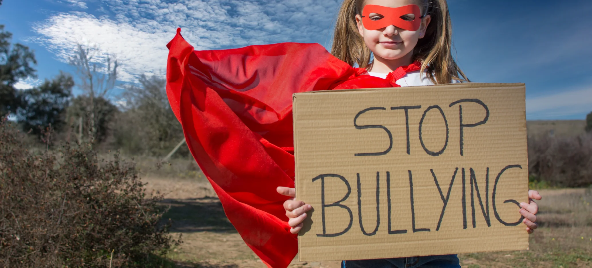 A child wearing a red mask and cape holds a cardboard sign that says “Stop bullying”.