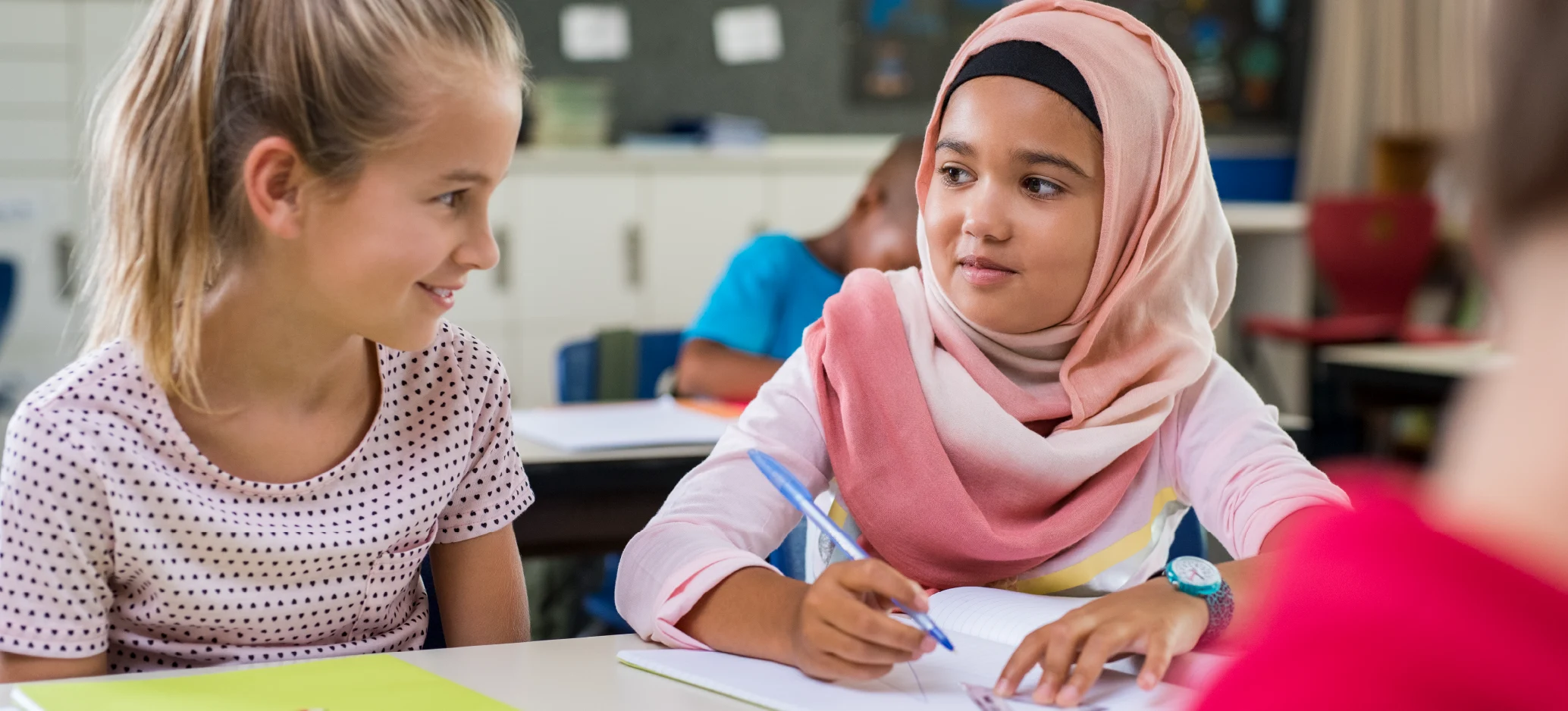 Two primary school girls sitting together at a desk, talking while one writes in a notebook.