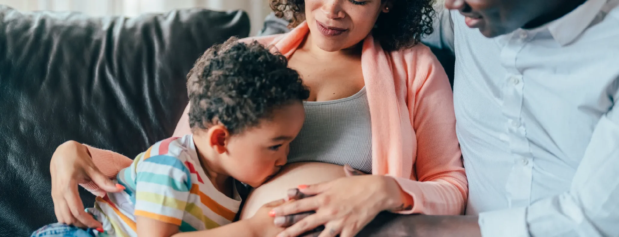 Toddler kissing pregnant mother’s belly while parents sit together on a sofa.