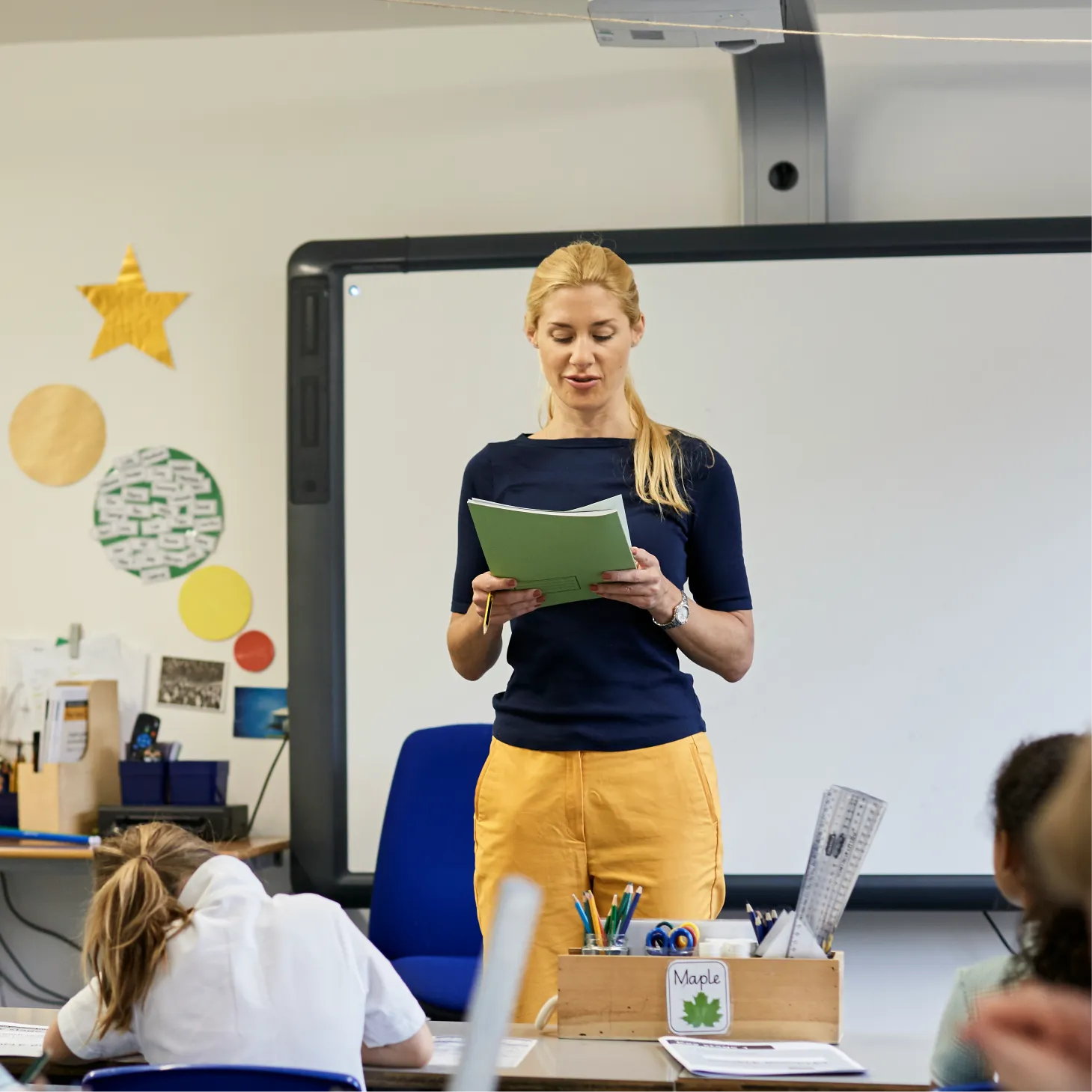 Teacher reading from an extract to the rest of her class, while they are heads-down at their desks writing.