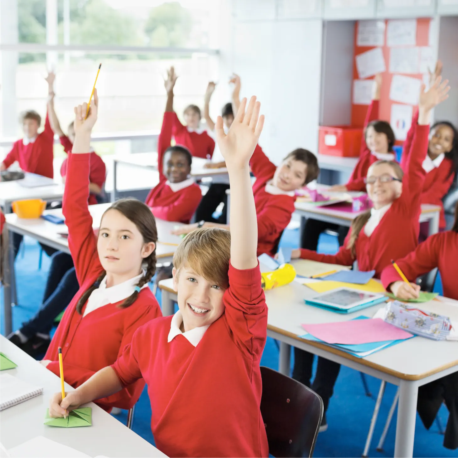 Rows of children in uniform at their tables, with their hands up looking keen to provide an answer.