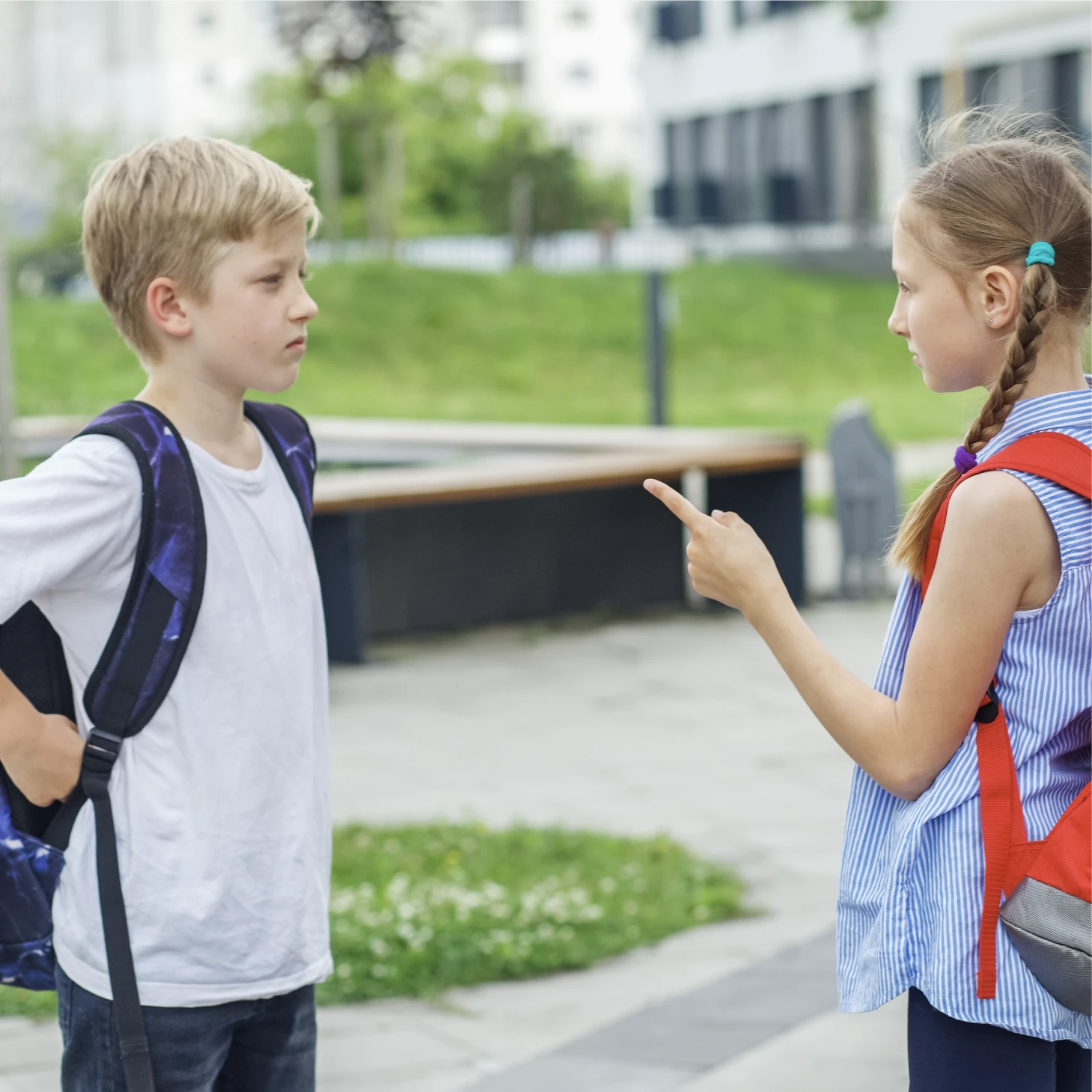 A girl points her finger at a boy as they face each other outdoors, both wearing school backpacks.