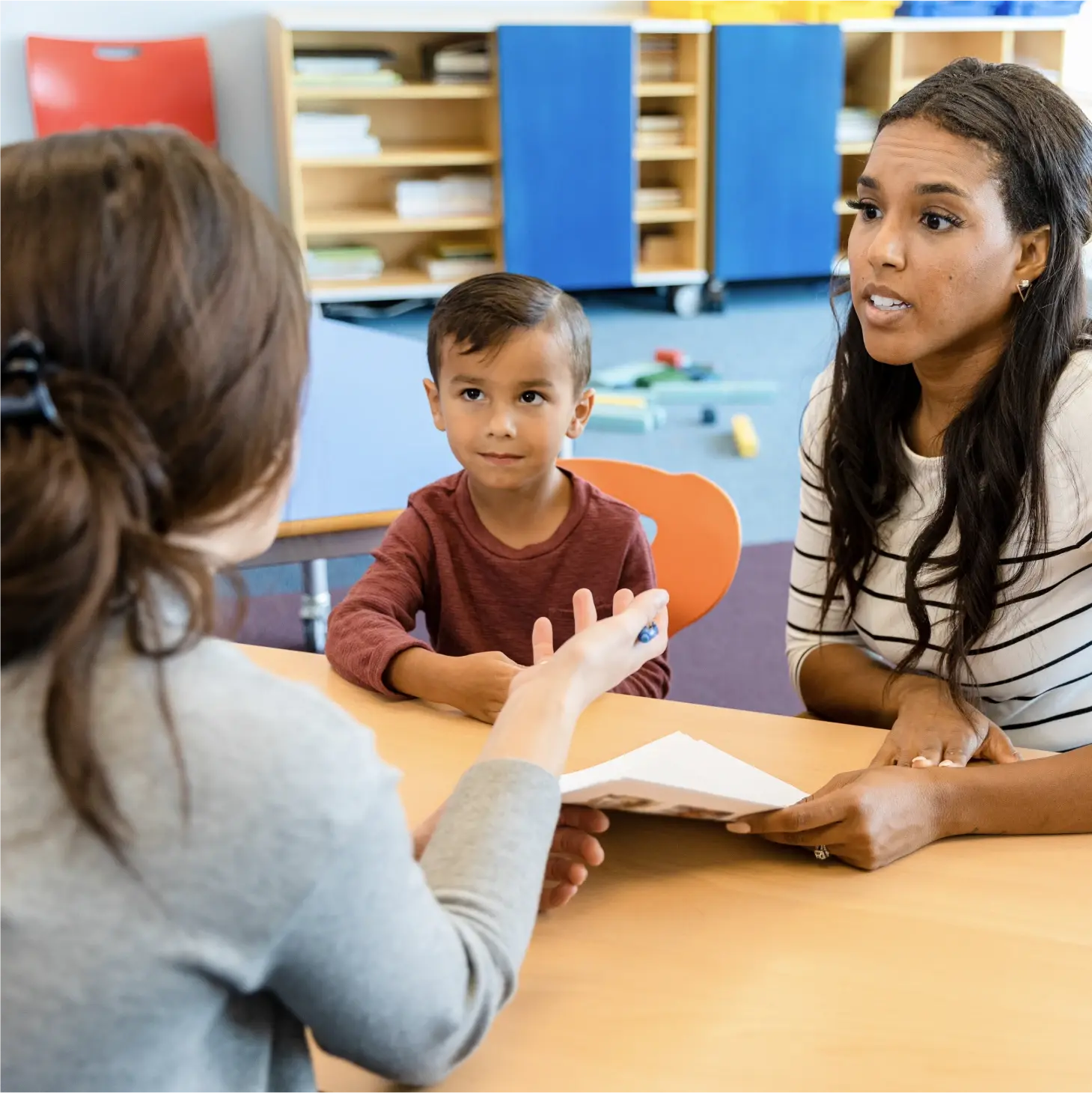 Teacher speaking with a parent and a young child at a table in a classroom.